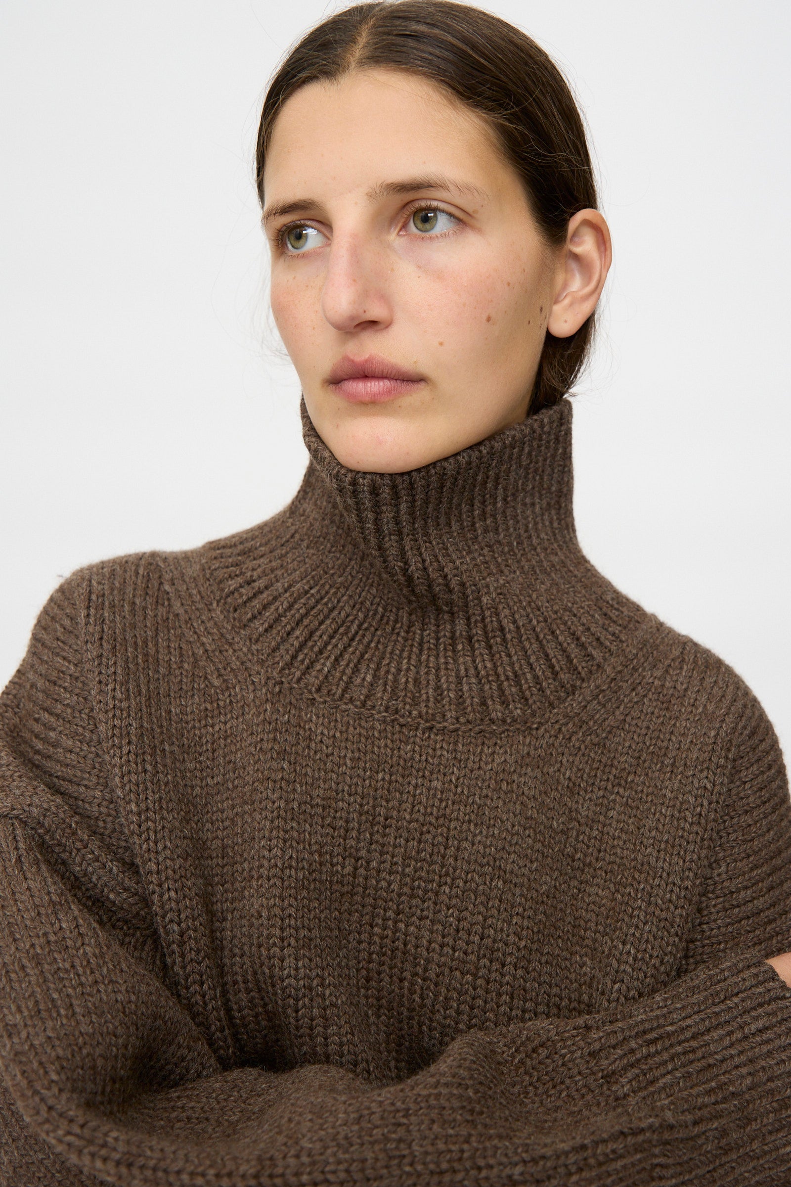 A person wears the Misha & Puff Turtleneck Sweater in Oak, featuring an oversized fit, thick highland wool, and a brown hue, while looking slightly to the side against a plain background.