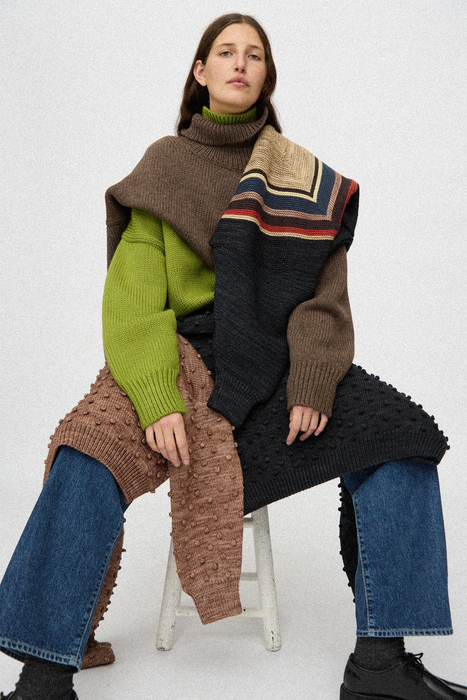 A woman sitting on a stool wearing Misha & Puff sweaters against a white background.