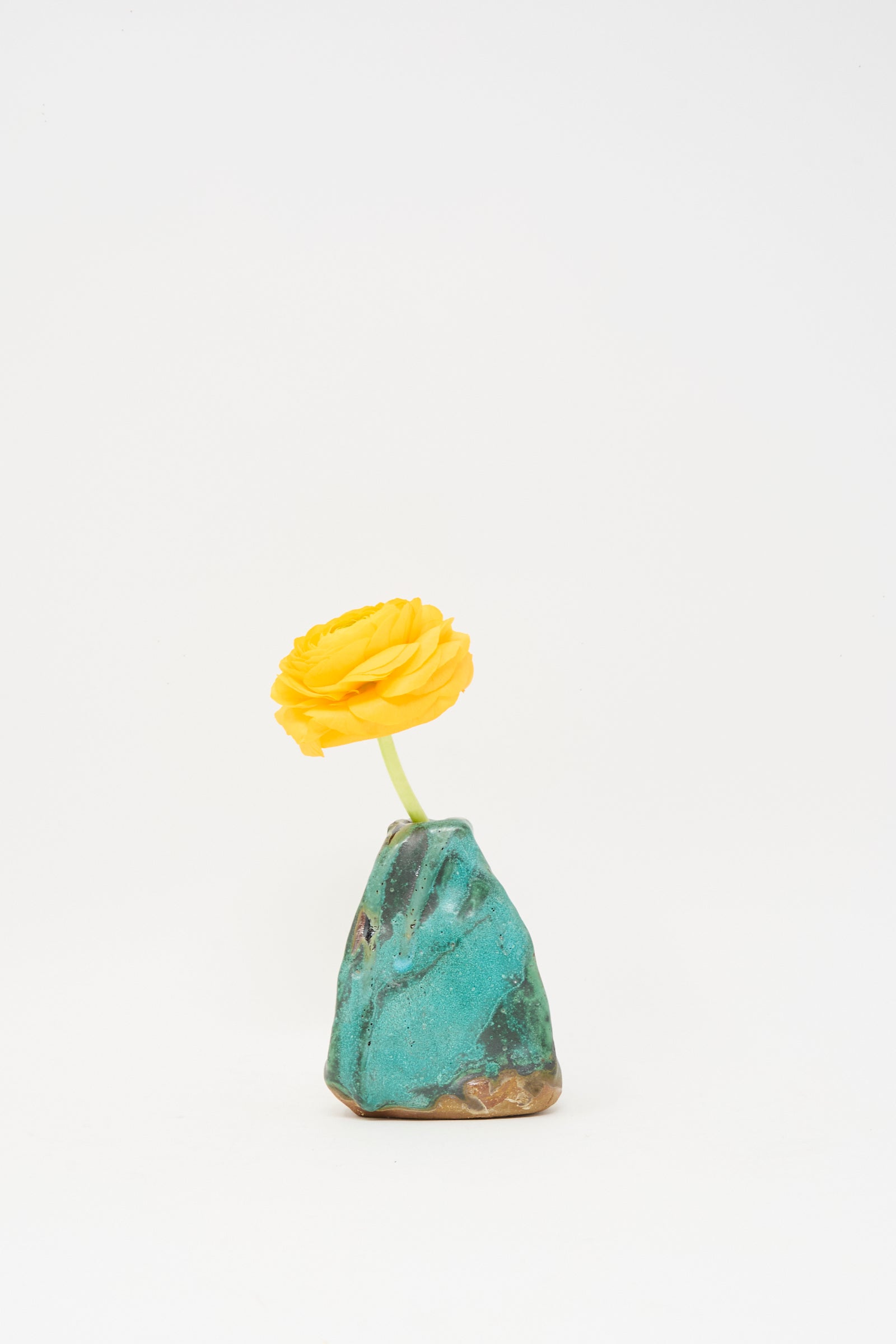 A single yellow flower sits in the Carved Bud Vase III by MONDAYS—a small, uniquely shaped turquoise and brown clay piece handcrafted in Brooklyn—set against a plain white background.