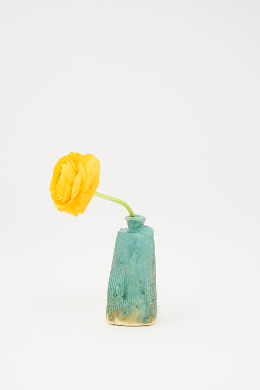 A single yellow flower with a green stem rests in the Carved Bud Vase IV by MONDAYS, a small turquoise vase with a ribbed pattern, crafted in Brooklyn and set against a plain white background.