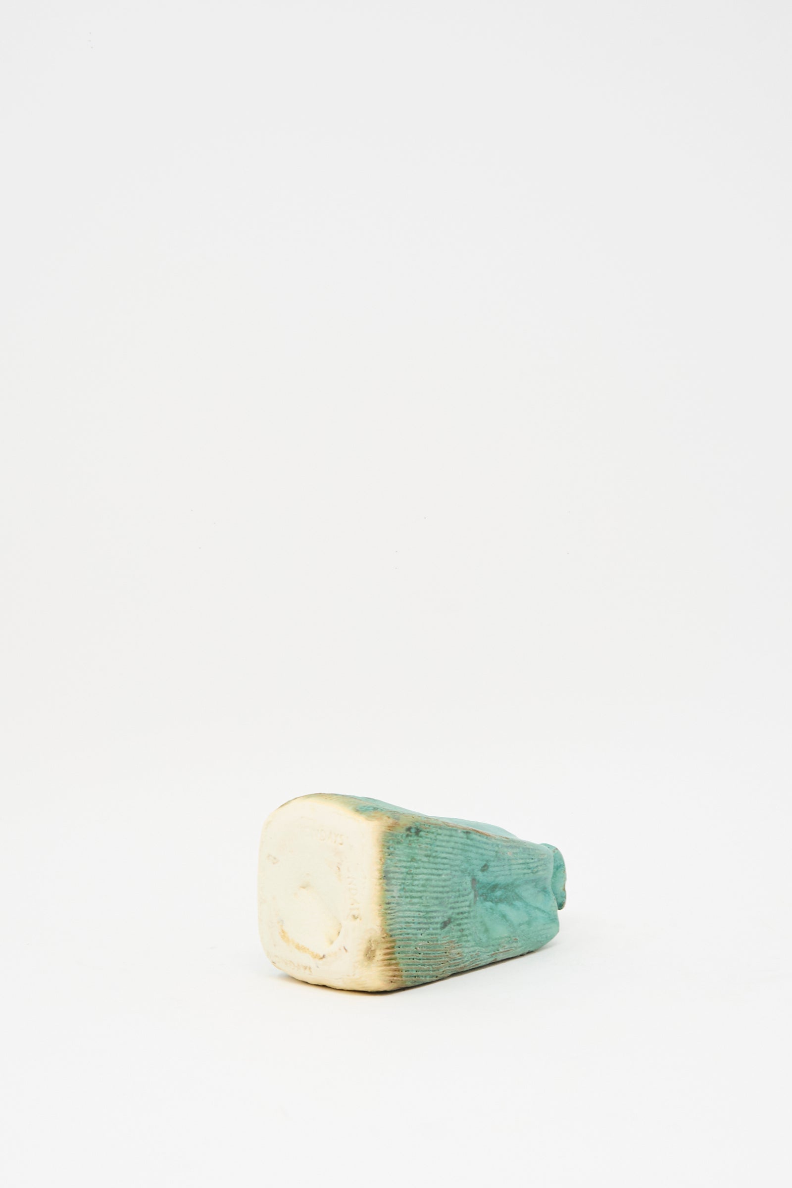 The Carved Bud Vase IV by MONDAYS, a small teal-glazed ceramic vase with a rectangular base and subtle rib pattern, rests on its side against a plain white background.