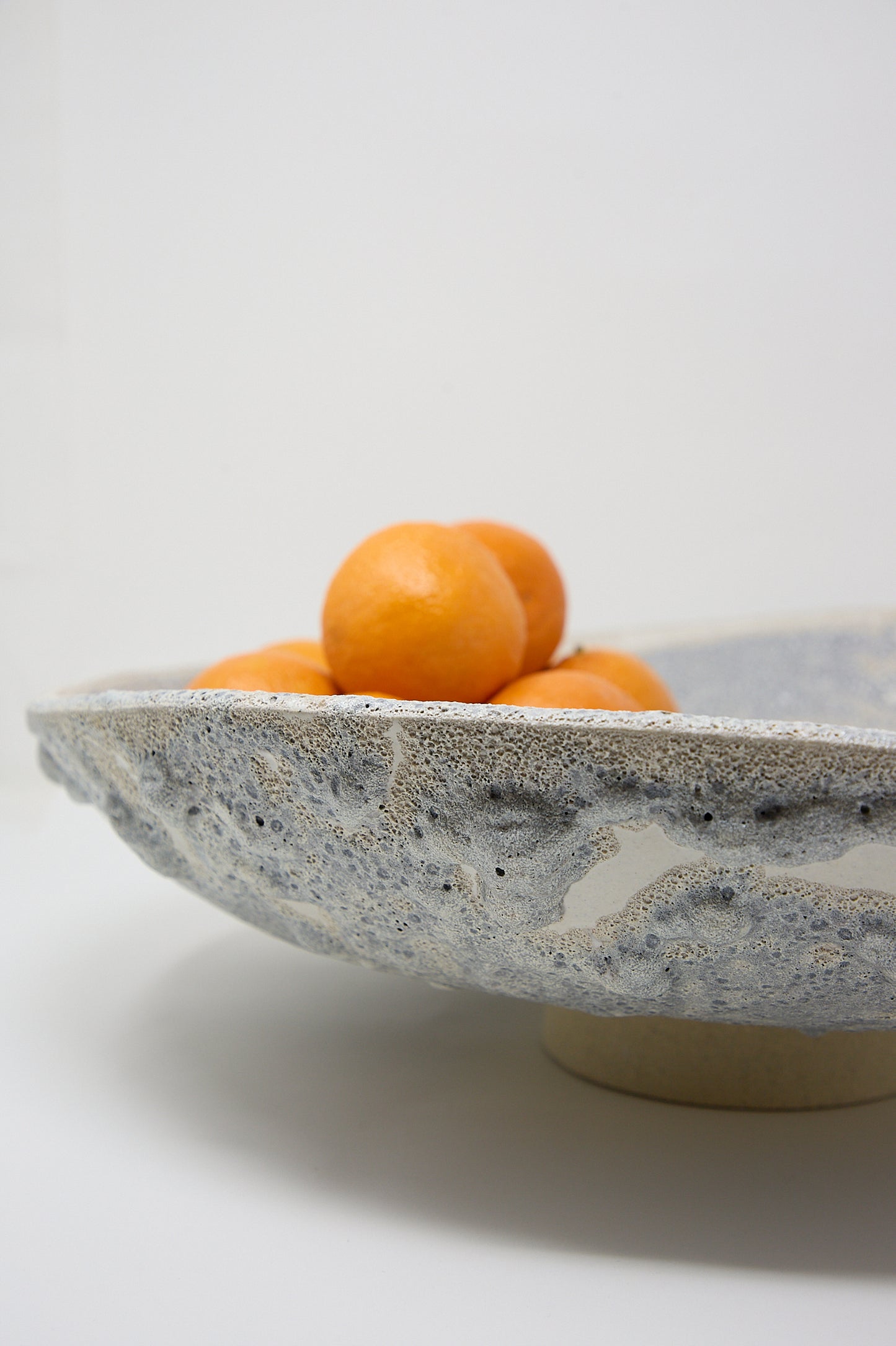 A hand-built Lava Pedestal Bowl in Glazed Stoneware by MONDAYS cradles several oranges against a plain white background.