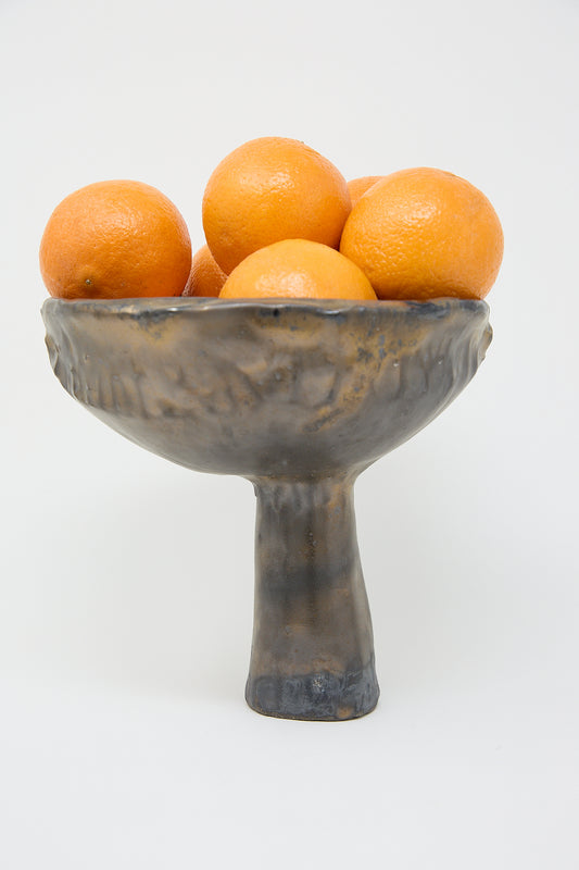 A MONDAYS Pedestal Vase in Bronze Glazed Stoneware with oranges in it, sitting on a pedestal.