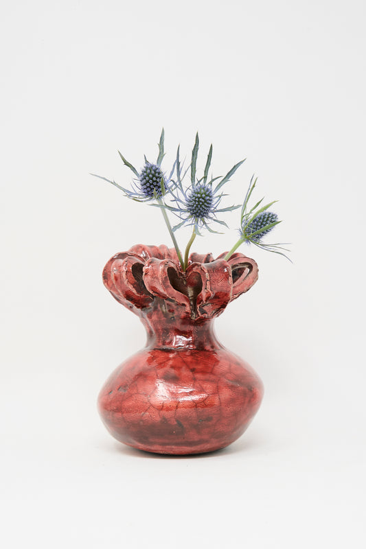 The MONDAYS Raku Ribbon Vase in Red, featuring a glossy finish and irregular ruffled rim, holds two spiky blue thistle blooms against a plain white background.