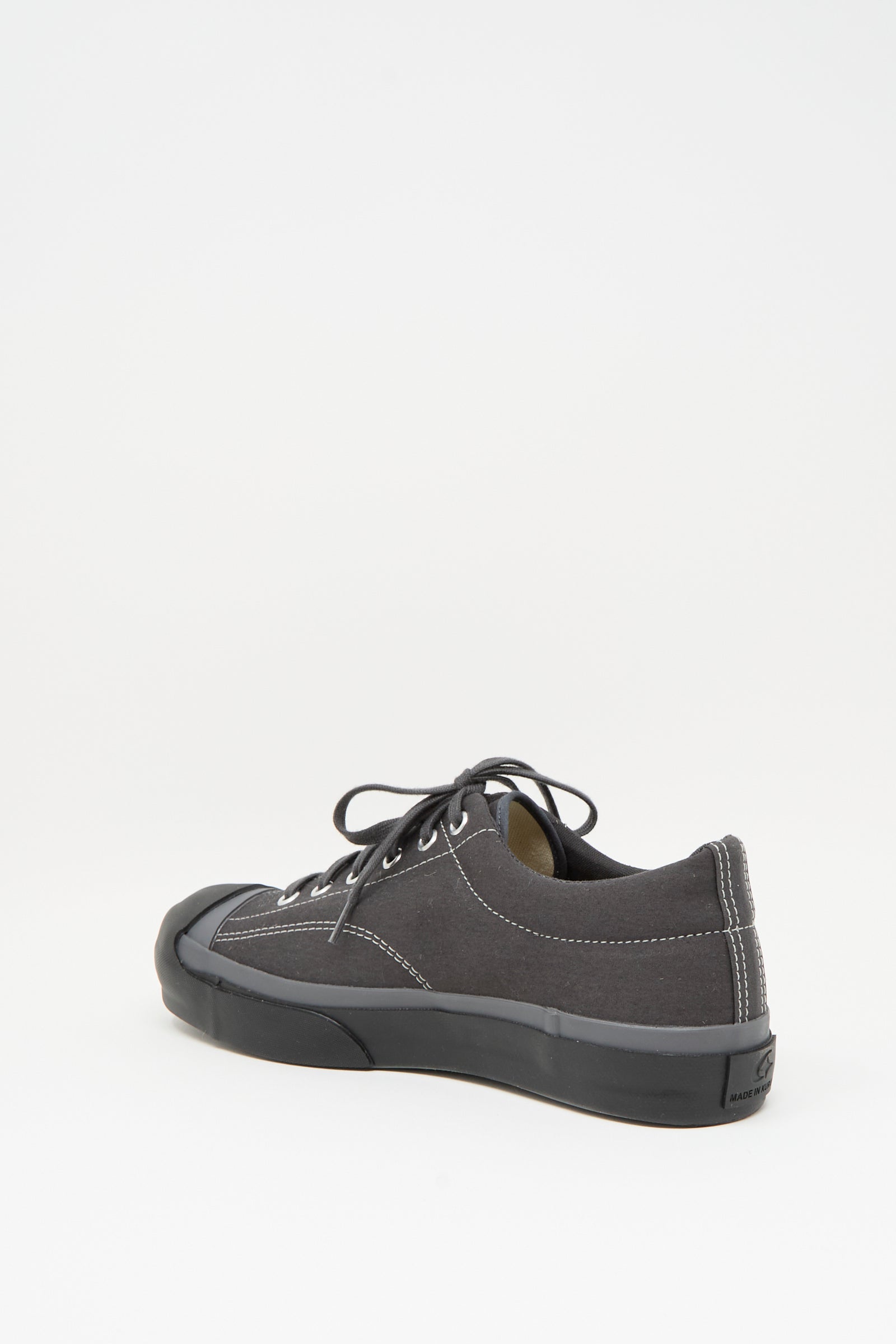 The Moonstar Gym Court Sneaker in Charcoal is a black low-top crafted from cotton nylon canvas, featuring black laces and white stitching, shown from the back against a plain white background.