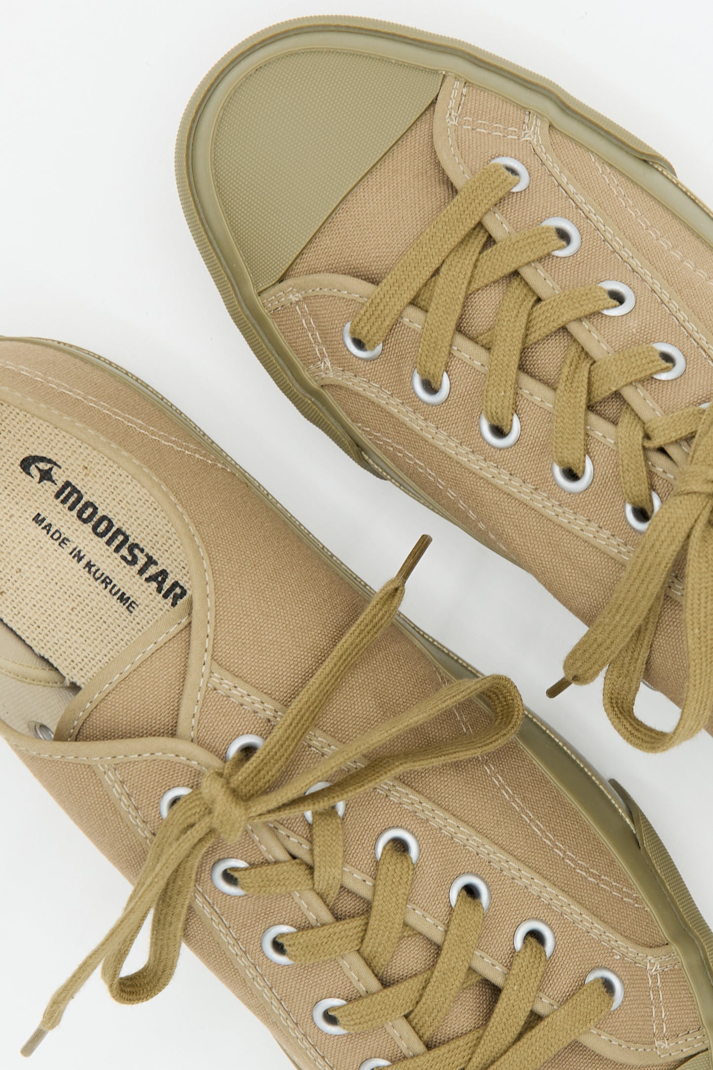 A pair of Low Trainer Sneaker in Khaki with beige rubber soles, laces, and metal eyelets. One shoe displays the brand "Moonstar" and "Made in Kurume" on the side. These unisex low trainers are handmade in Japan, combining traditional craftsmanship with modern style.