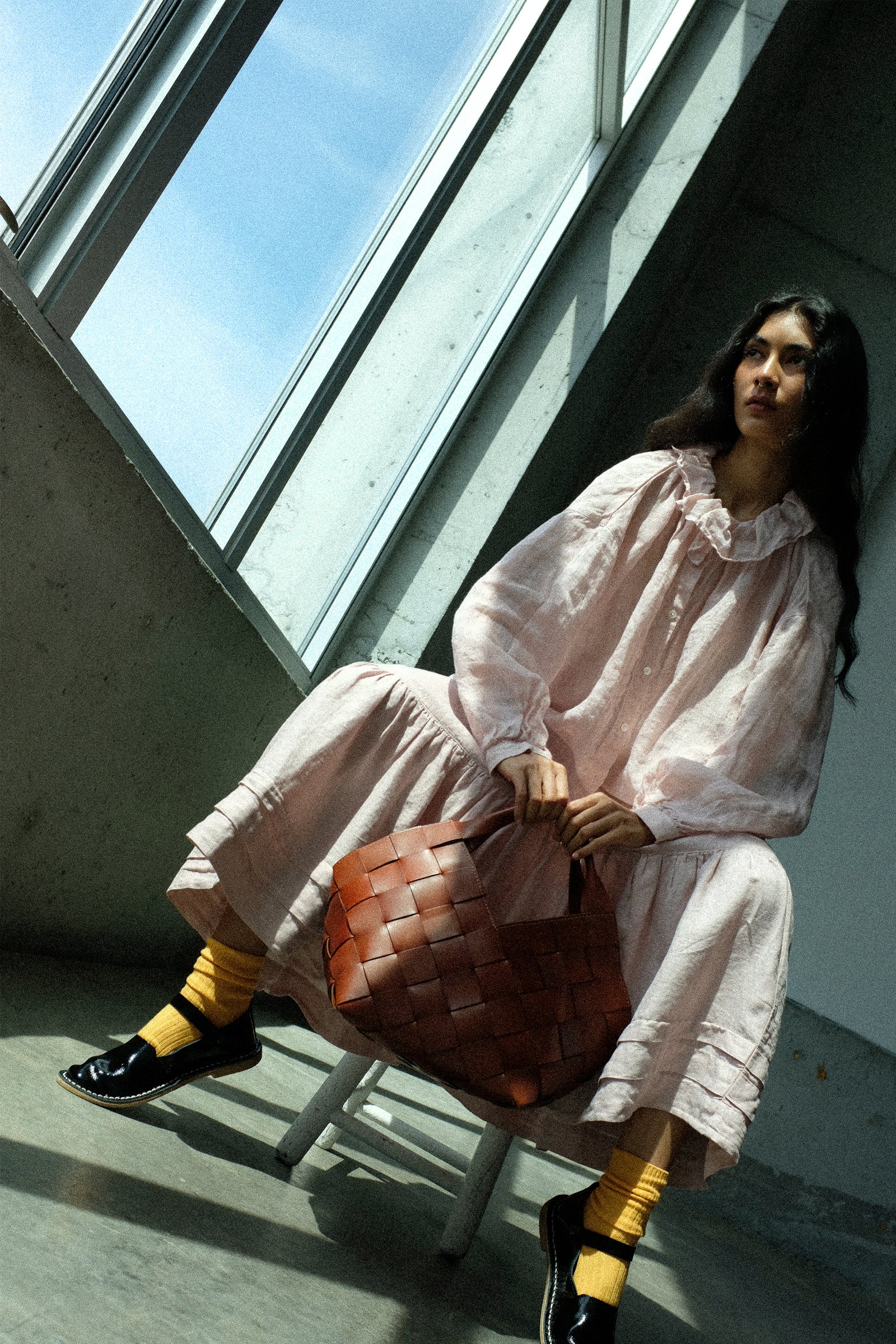 Woman in wearing nest Robe's Linen Gauze Frill Blouse in Light Peach, and Linen Natural Dyed Pleated Gathered Skirt in Pink, sitting by a large window holding the Dragon Diffusion Botteghe Bag in Tan.