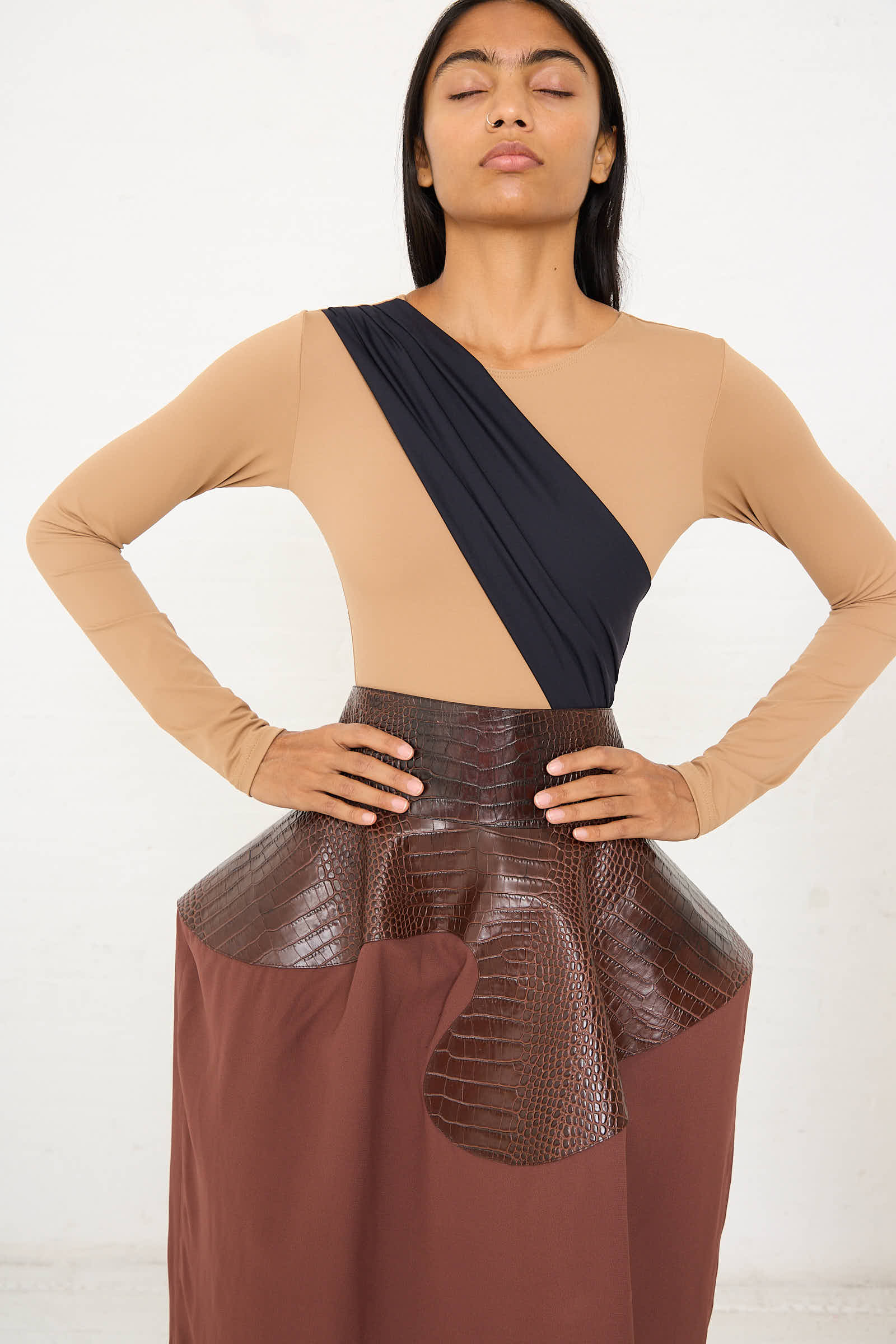 A person stands with eyes closed, wearing the Alix Sleeved Bodysuit in Black and Nude by Niccolò Pasqualetti, paired with a brown skirt featuring a textured, glossy section. Their hands rest on their hips against a plain light background.