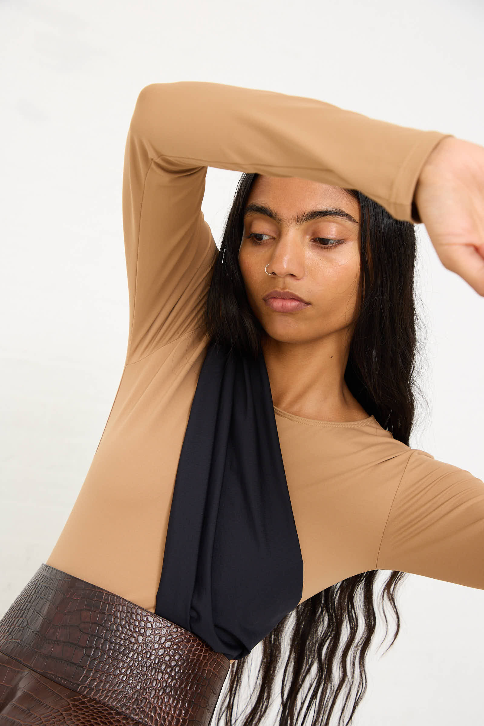 Person with long dark hair wearing the Niccolò Pasqualetti Alix Sleeved Bodysuit in Nude, complemented by a black scarf and a brown textured skirt, posing with arms raised against a plain white background. The slim fit stretch lycra bodysuit accentuates the elegant simplicity of the outfit.