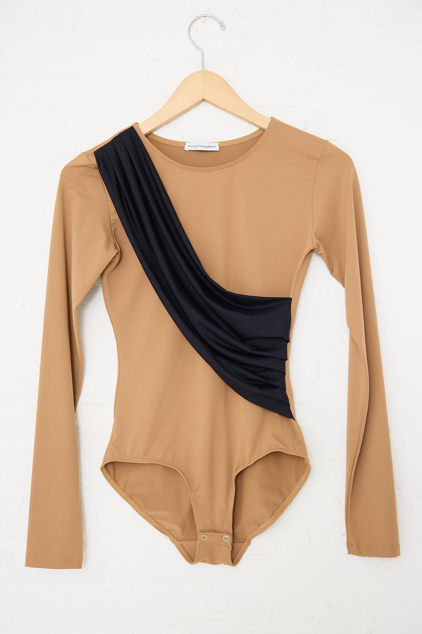 The Alix Sleeved Bodysuit in Black and Nude by Niccolò Pasqualetti features a slim fit, beige long-sleeves, and an asymmetrical black draped detail on one side. It is crafted from stretch lycra and is shown hanging on a wooden hanger against a white background.