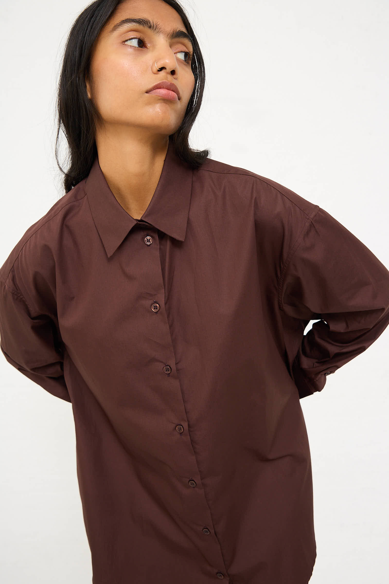 A person in a Niccolò Pasqualetti Cotton Poplin Classico Shirt in Brown with an oversized fit and long sleeves stands against a plain background, looking slightly to the side.