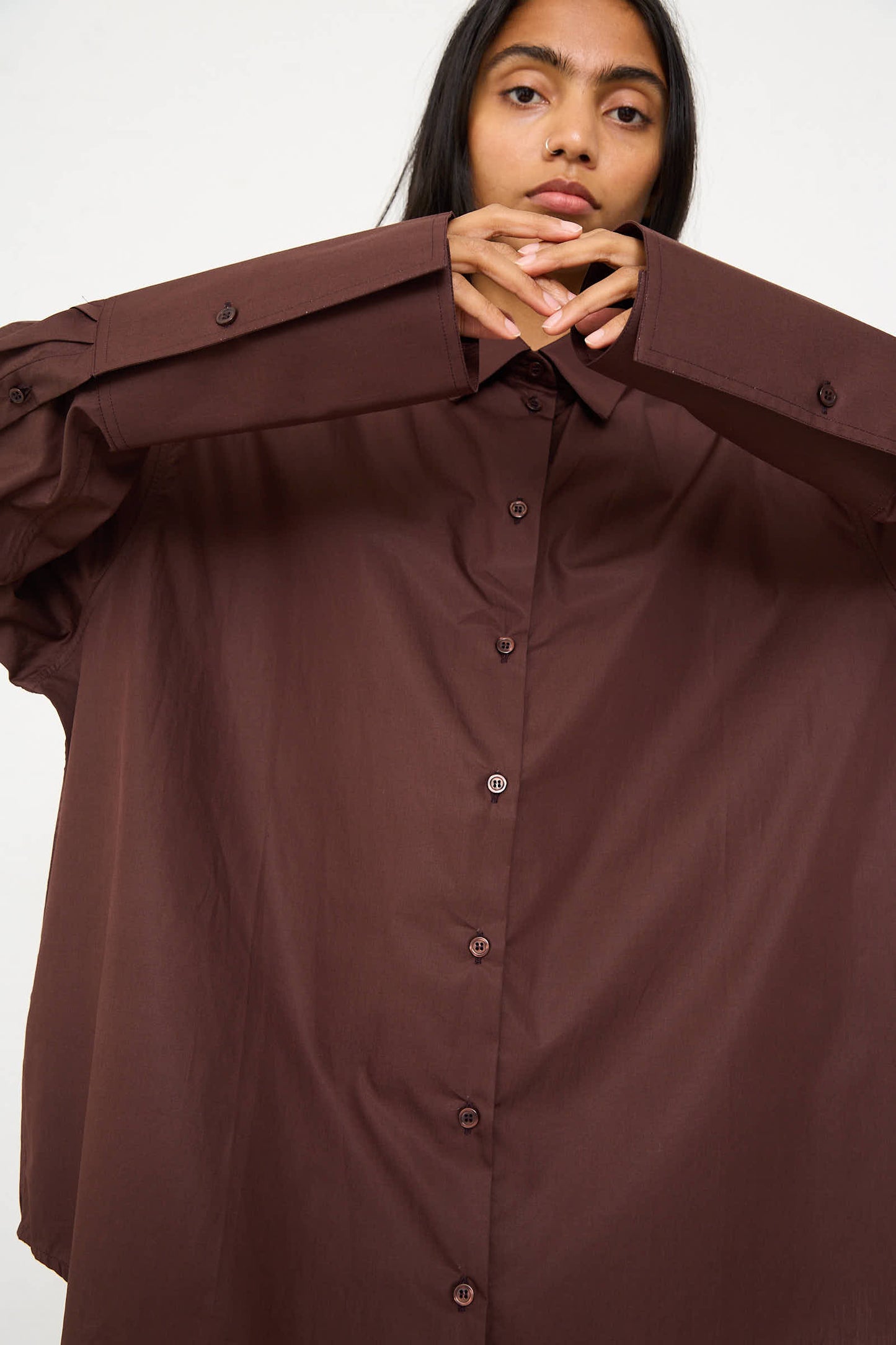 A person with long dark hair wearing the Niccolò Pasqualetti Cotton Poplin Classico Shirt in Brown looks directly at the camera with their hands raised near their face.