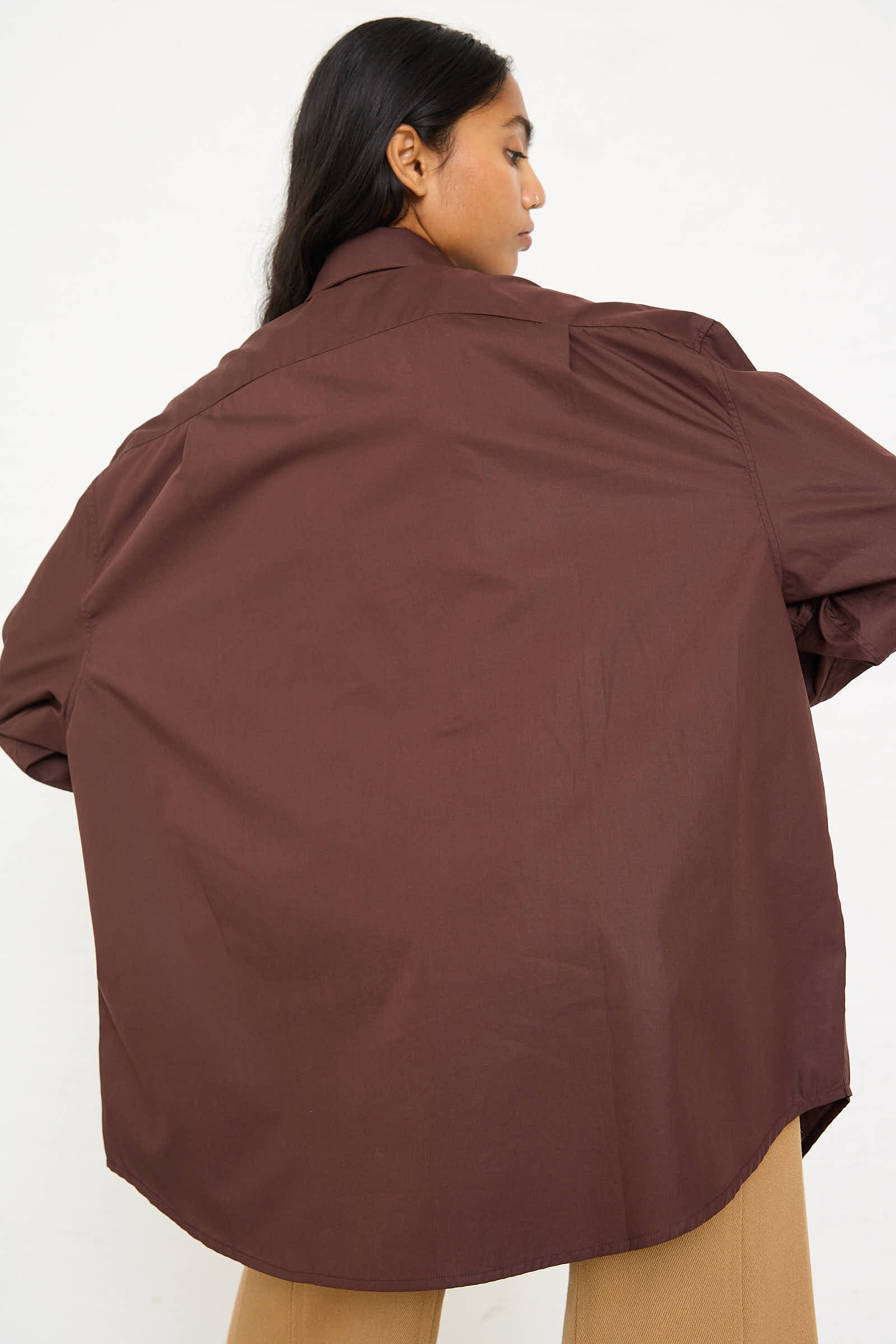 A person with long hair, seen from the back wearing an oversized Niccolò Pasqualetti Cotton Poplin Classico Shirt in Brown and light brown pants, poses against a plain background.