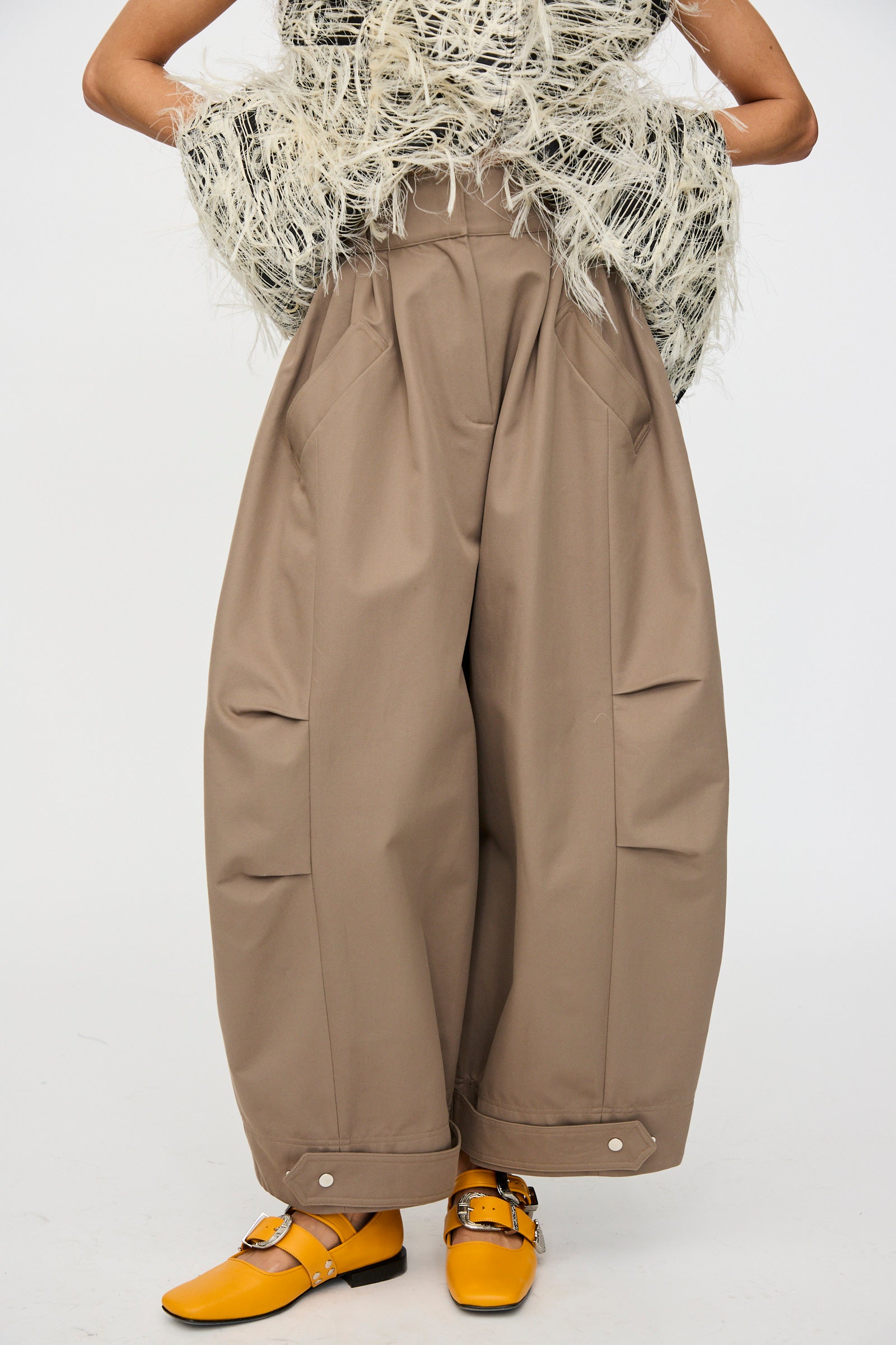 A person wears Niccolò Pasqualetti’s Cotton Twill Montage Trousers in Brown, paired with a textured off-white top and yellow buckle shoes, standing against a plain background.