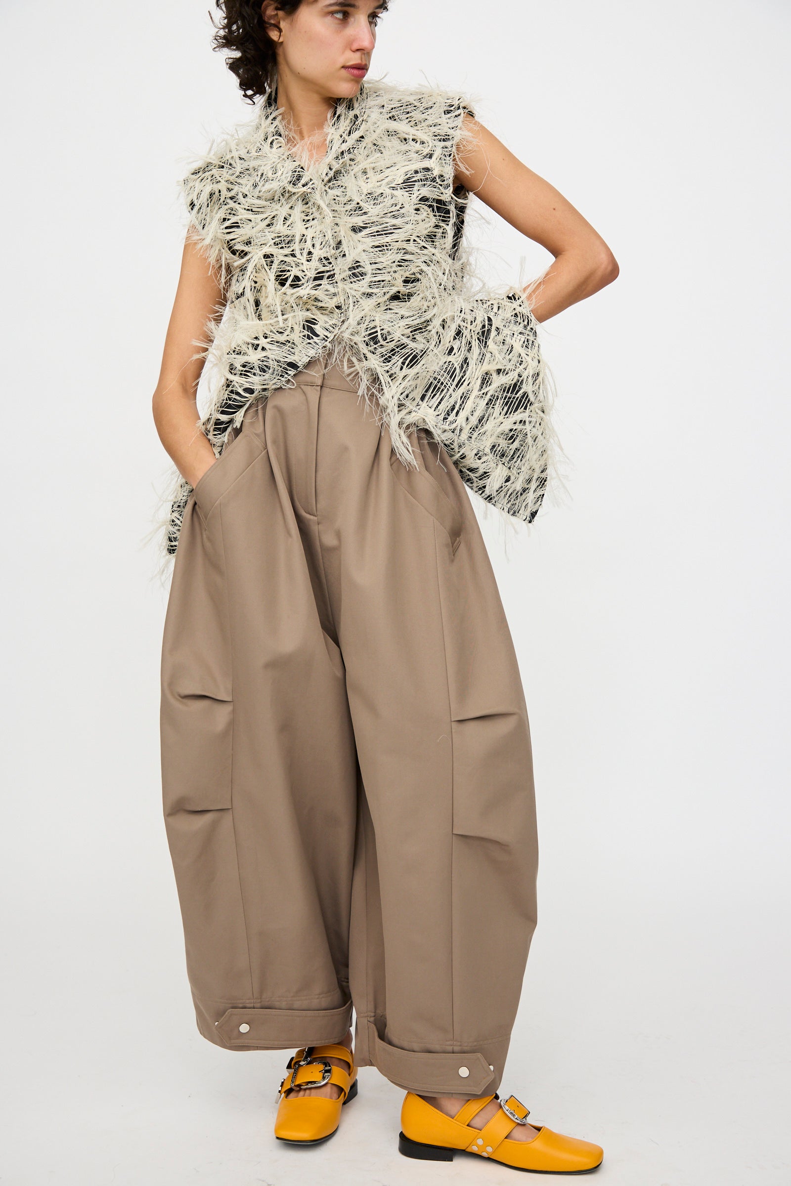A person wears Niccolò Pasqualetti’s Cotton Twill Montage Trousers in Brown with a textured sleeveless vest and yellow Mary Jane shoes, standing against a plain background.