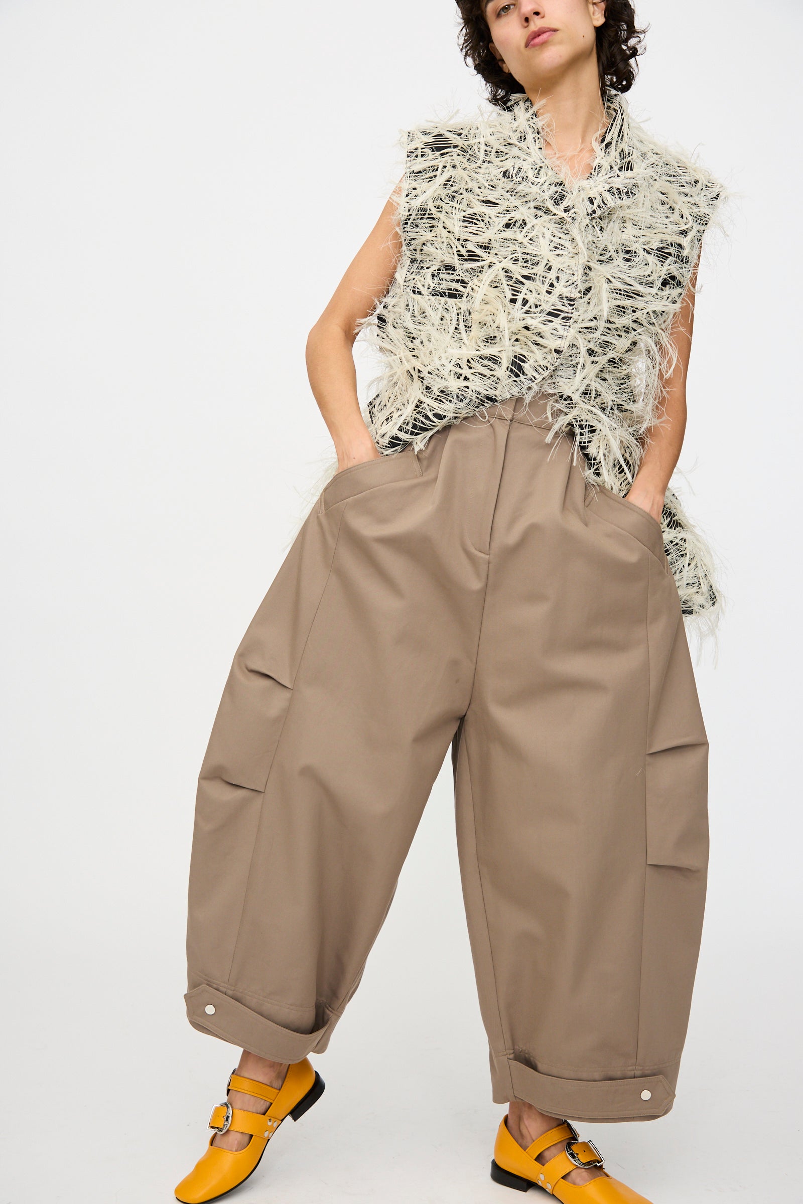 A person stands with hands in pockets against a plain background, wearing a sleeveless textured top, Niccolò Pasqualetti Cotton Twill Montage Trousers in Brown, and yellow strap shoes.