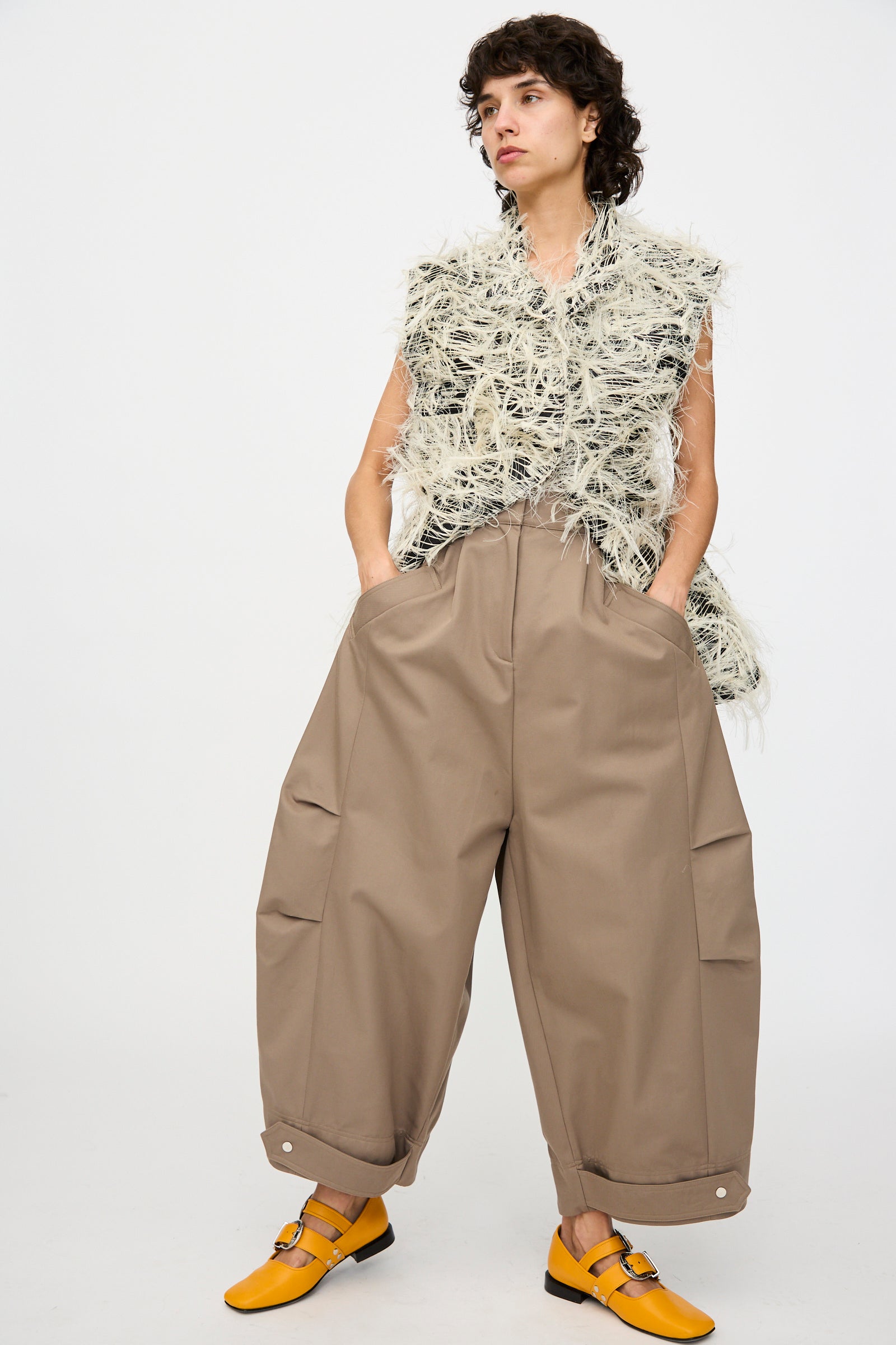 A person stands hands-in-pockets against a plain backdrop, wearing Niccolò Pasqualetti's Cotton Twill Montage Trousers in Brown—high-waisted, oversized with wide legs—paired with a sleeveless textured top and yellow shoes.