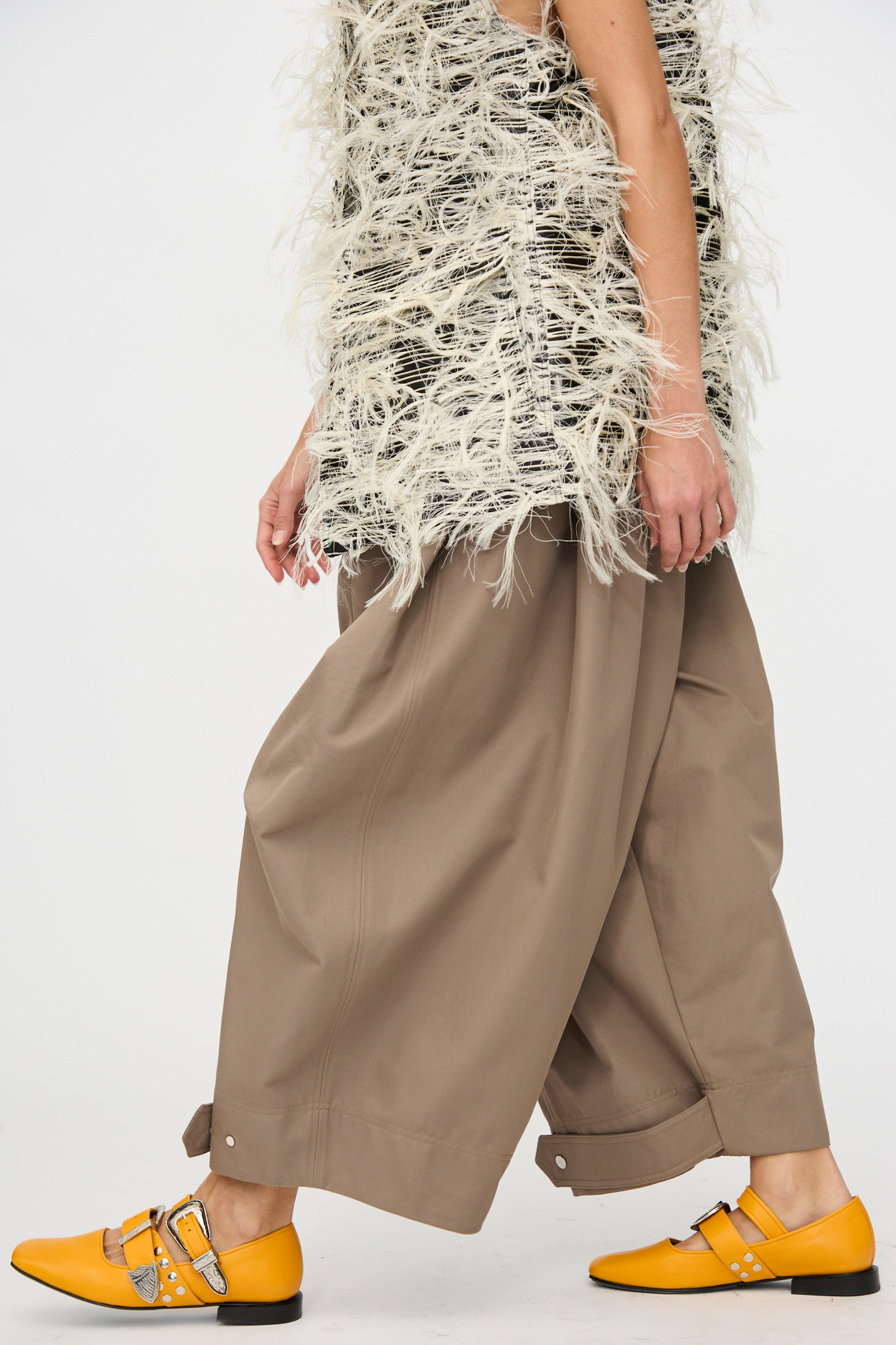 A person in Cotton Twill Montage Trousers in Brown by Niccolò Pasqualetti, a sleeveless fringed top, and bright yellow buckle shoes walks against a plain white background.