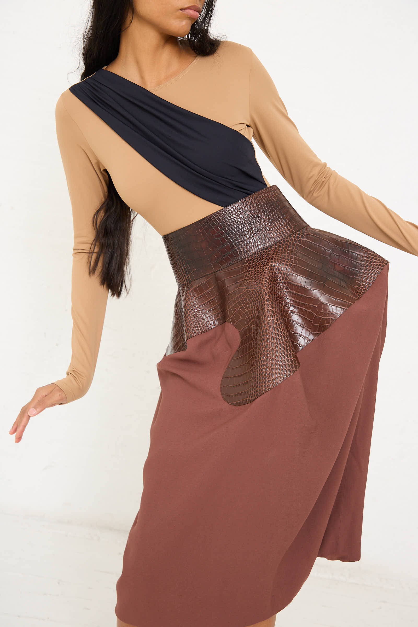 A person wearing a long-sleeve beige and black top with the Niccolò Pasqualetti Croc Embossed Leather and Viscose Crepe Onda Skirt in Brown and Burnt Brown poses against a plain white background.