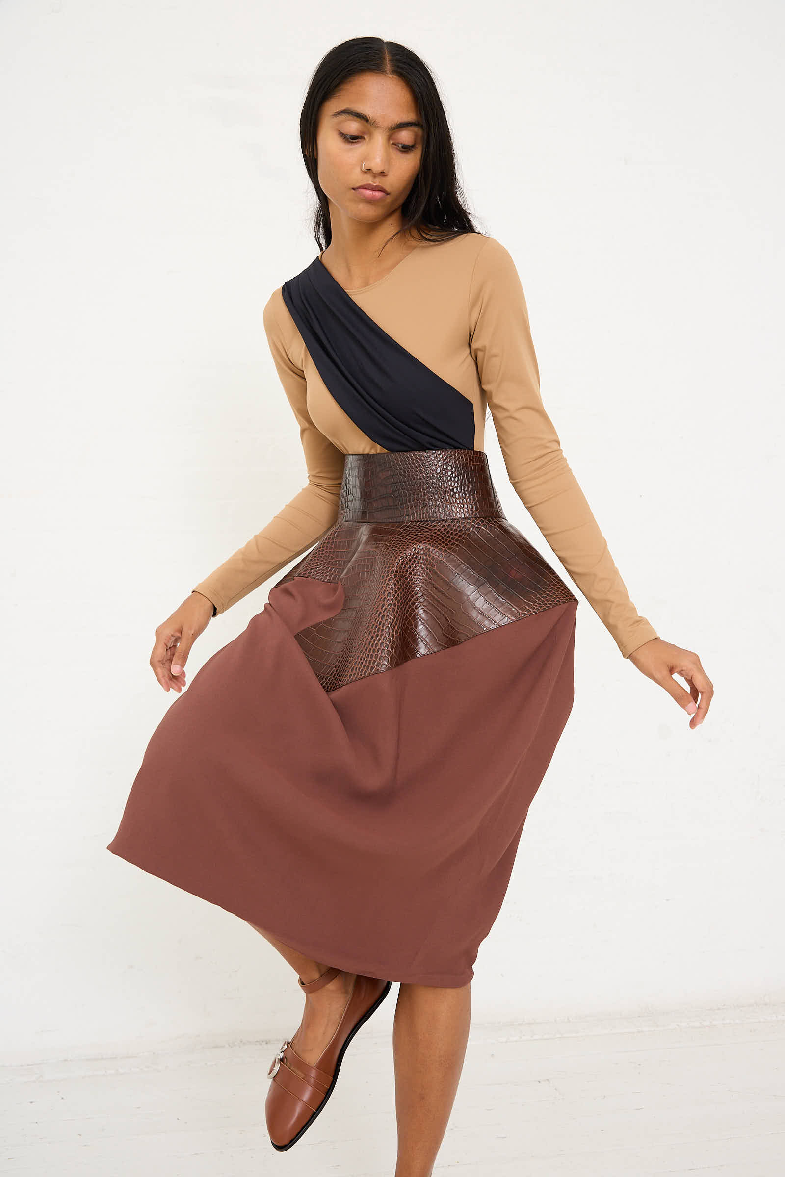 A woman in a beige and black long-sleeve top poses against a white background, wearing the Niccolò Pasqualetti Croc Embossed Leather and Viscose Crepe Onda Skirt in Brown and Burnt Brown, paired with brown shoes. She stands in a relaxed stance.