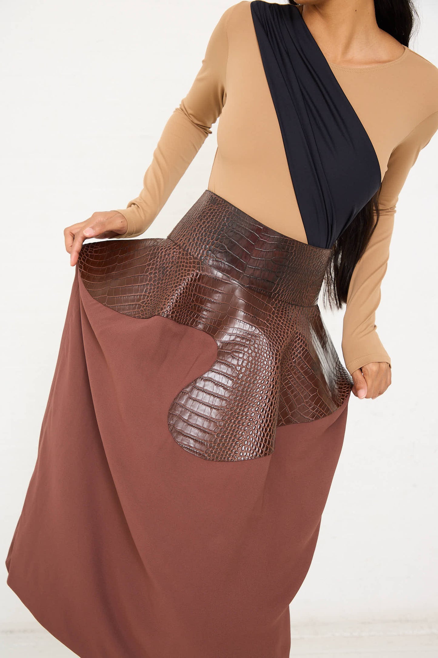 A person wearing a Niccolò Pasqualetti Croc Embossed Leather and Viscose Crepe Onda Skirt in Brown and Burnt Brown with a beige top and black accent, standing against a light background.