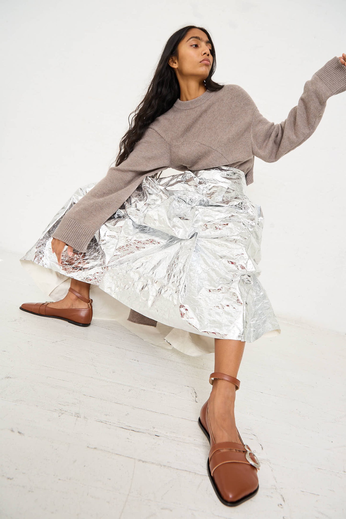 A woman wearing a beige sweater, the Papery Metallic Leather Lattina Skirt in Silver by Niccolò Pasqualetti with a deconstructed A-line silhouette, and brown flat shoes poses against a plain white background.