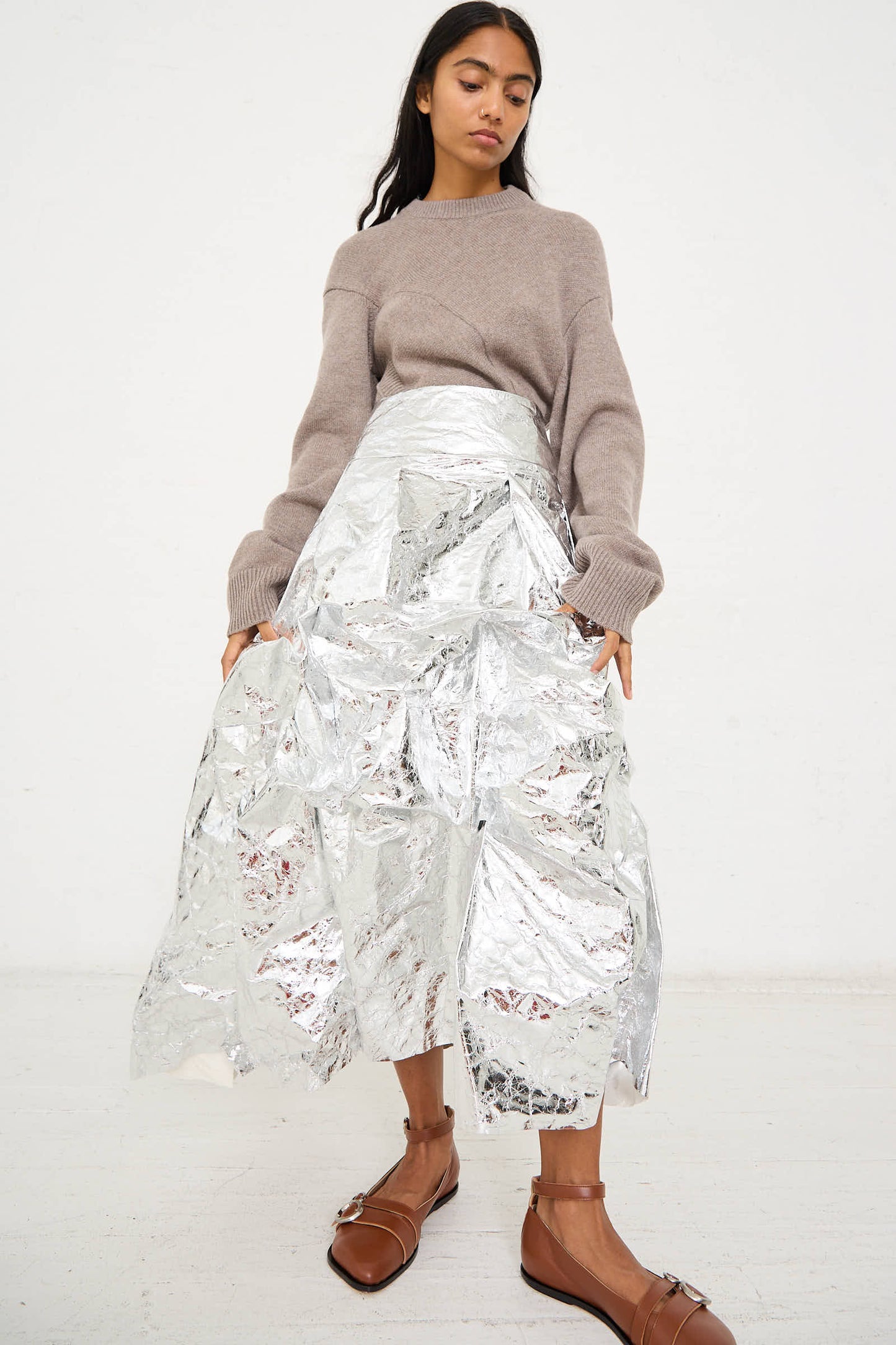 A person is wearing a beige sweater, Niccolò Pasqualetti's Papery Metallic Leather Lattina Skirt in Silver, and brown sandals while standing against a plain white background.