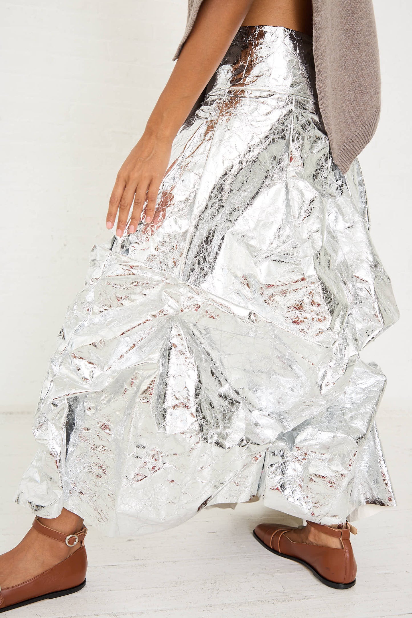 A person wearing a Niccolò Pasqualetti Papery Metallic Leather Lattina Skirt in Silver and brown flat shoes is walking indoors. Only their lower body is visible.