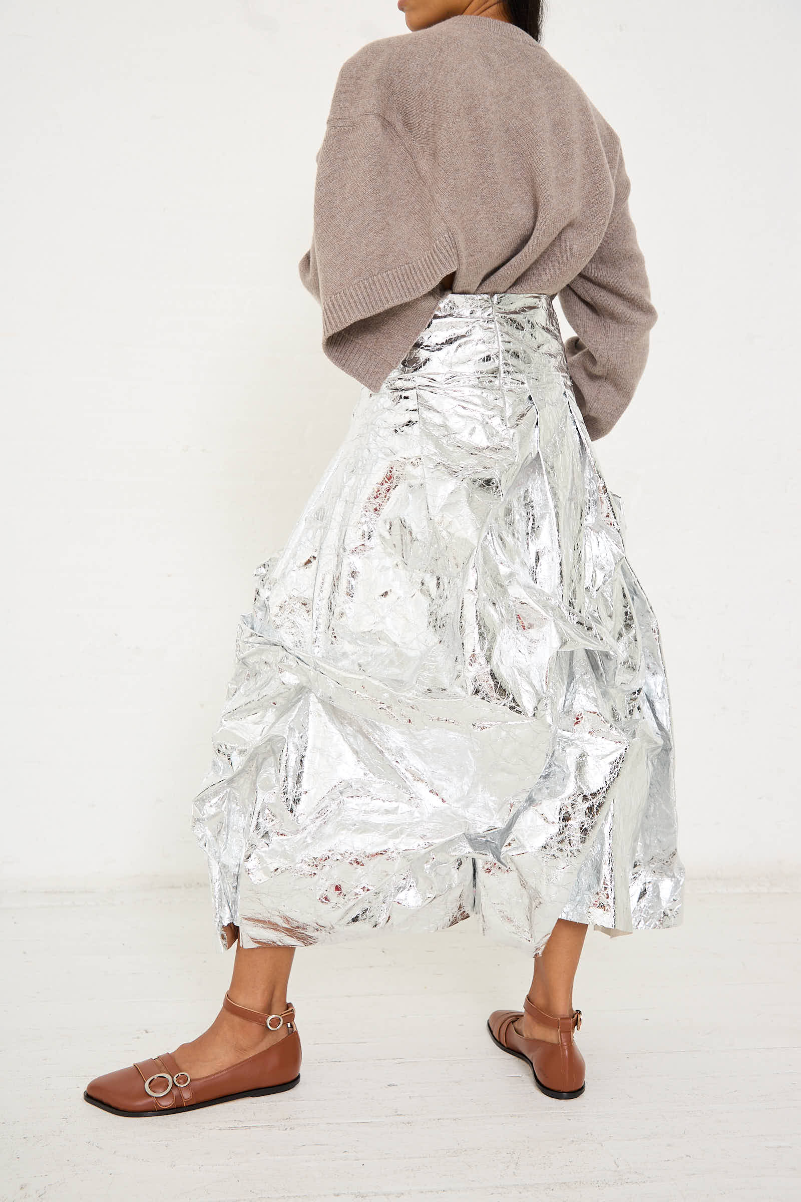 Person wearing a taupe sweater, the Niccolò Pasqualetti Papery Metallic Leather Lattina Skirt in Silver with a deconstructed A-line silhouette, and brown flats with buckles standing against a plain white background.