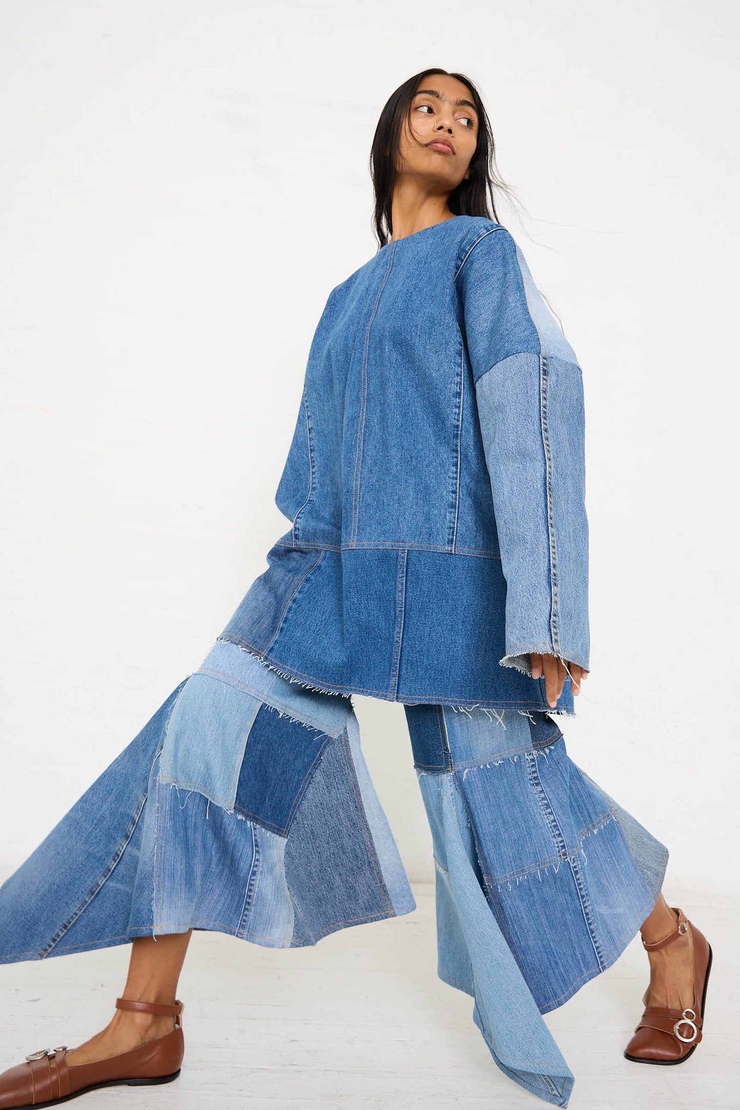 A person poses wearing a fashion ensemble made from various shades of denim. The outfit consists of a long-sleeved top and the Niccolò Pasqualetti Salvaged Jean Patchwork Lengo Trouser in Blue, which features mid-rise trousers with patchwork detailing and flared legs.