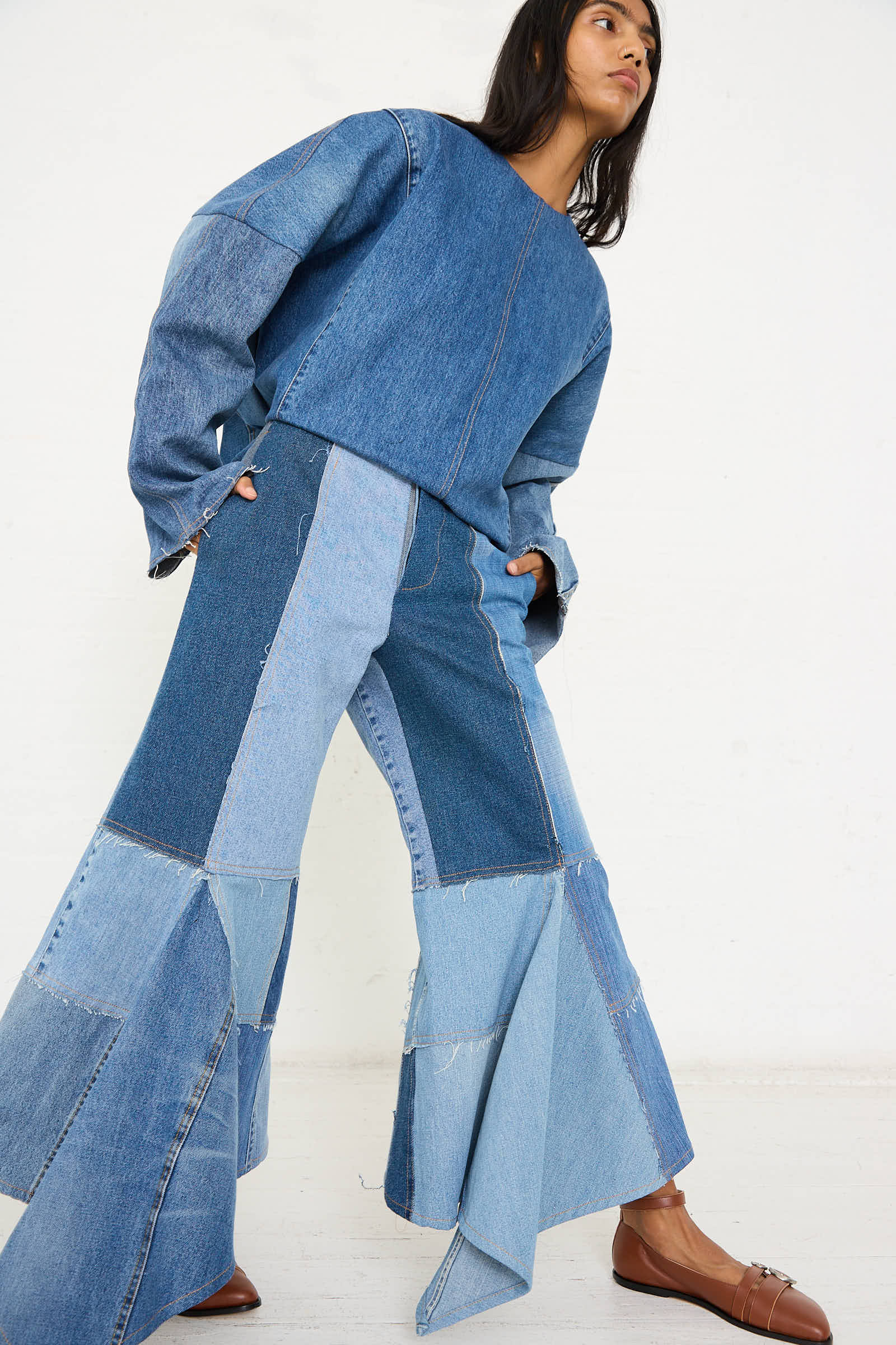 A person wearing Niccolò Pasqualetti's Salvaged Jean Patchwork Lengo Trousers in Blue with brown loafers, standing against a plain white backdrop.