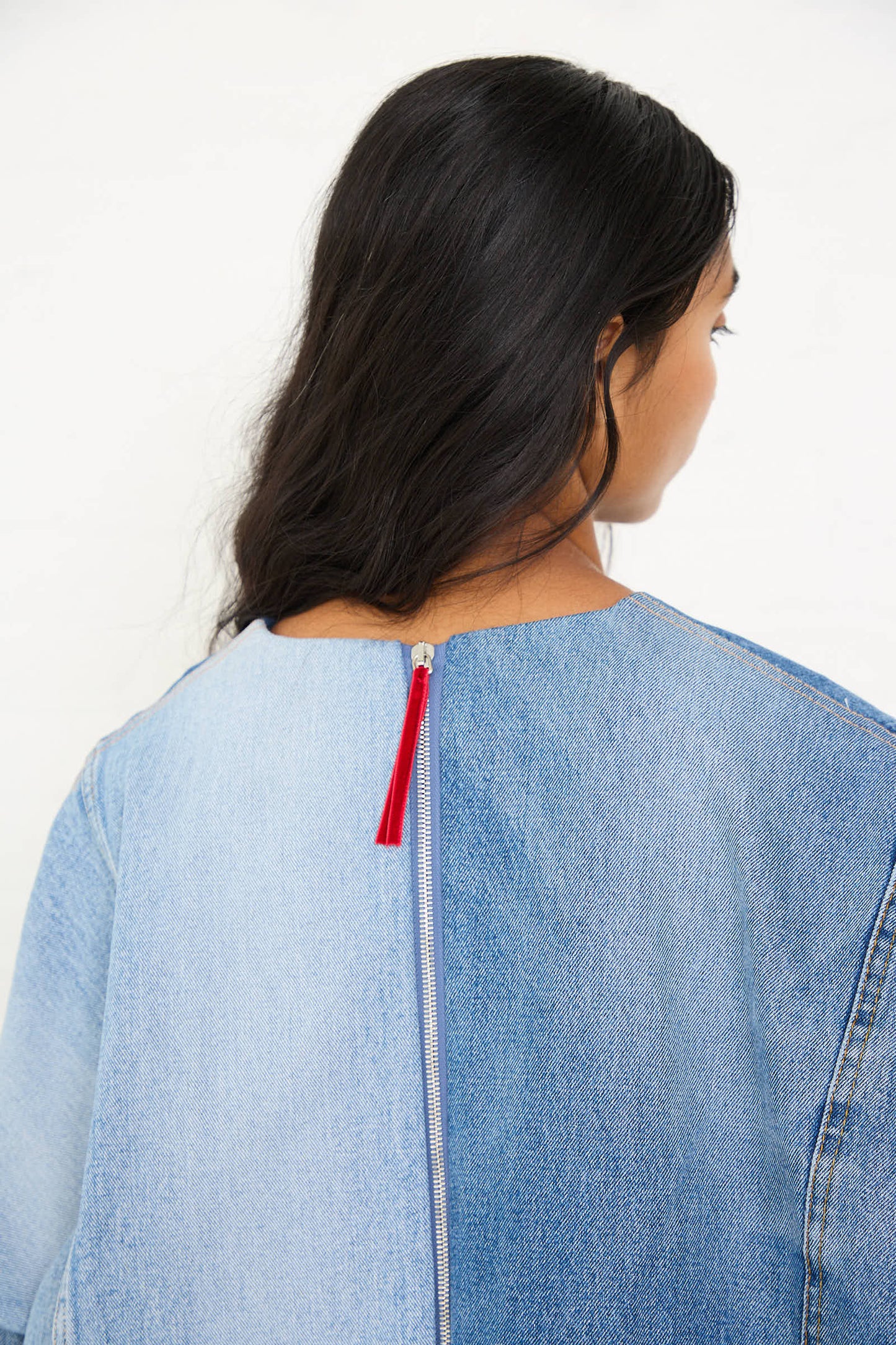 Revised Sentence: Rear view of a person with long dark hair wearing the oversized fit Salvaged Jean Patchwork Raglan Shirt in Blue by Niccolò Pasqualetti, featuring a red zipper pull at the back.