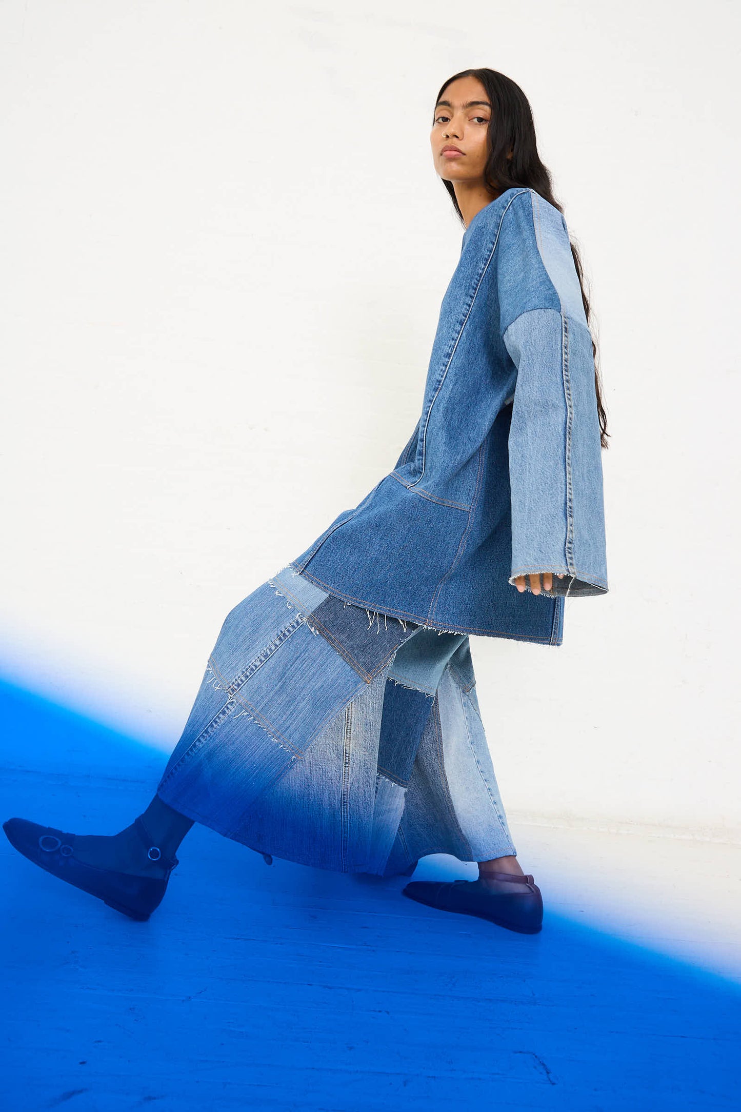 A person wearing the Salvaged Jean Patchwork Raglan Shirt in Blue by Niccolò Pasqualetti and dark shoes is walking in front of a white and blue gradient background, their long sleeve top billowing gracefully.