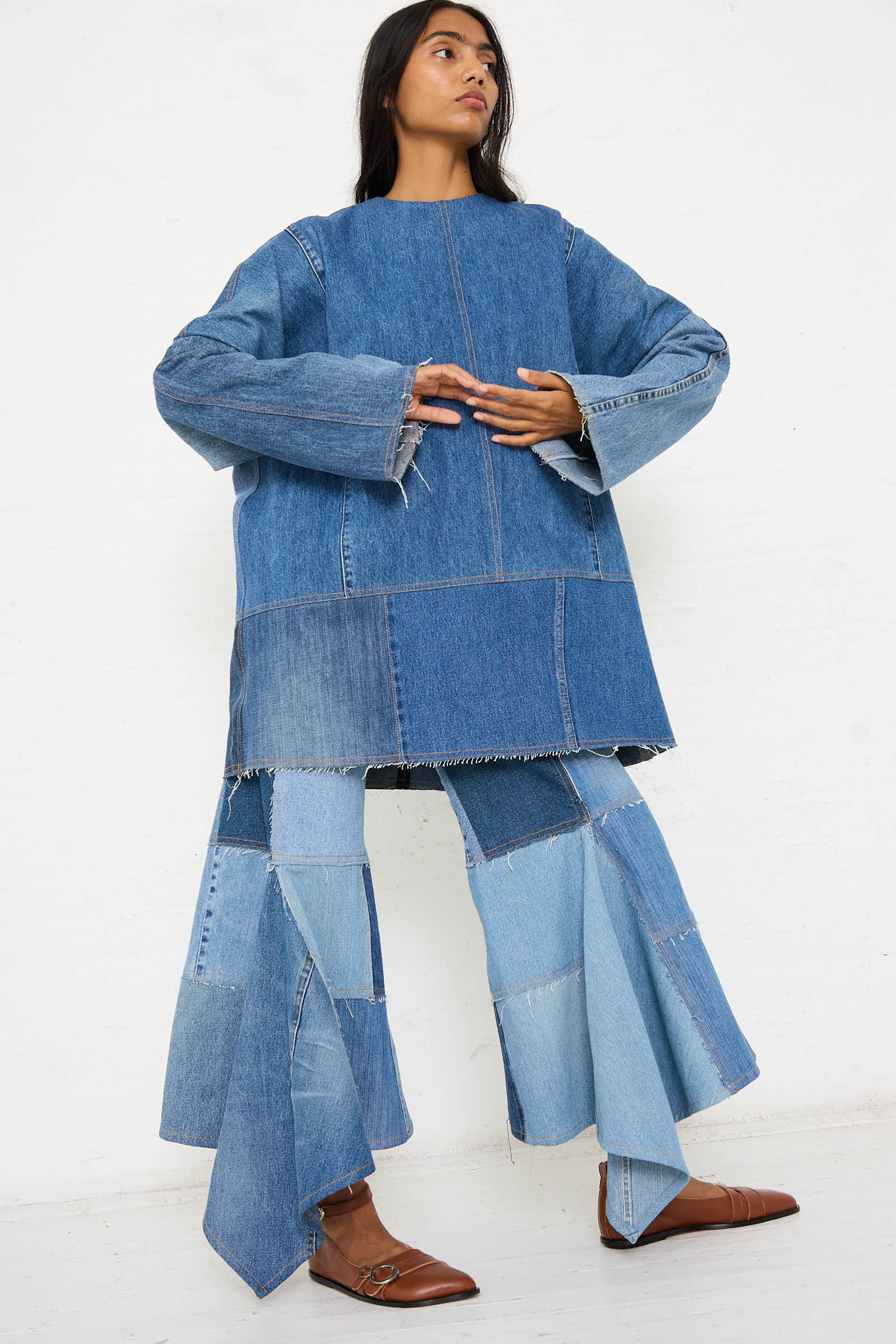 Person wearing a Salvaged Jean Patchwork Raglan Shirt in Blue by Niccolò Pasqualetti, along with wide-legged pants. They stand against a plain white background and look to the side.