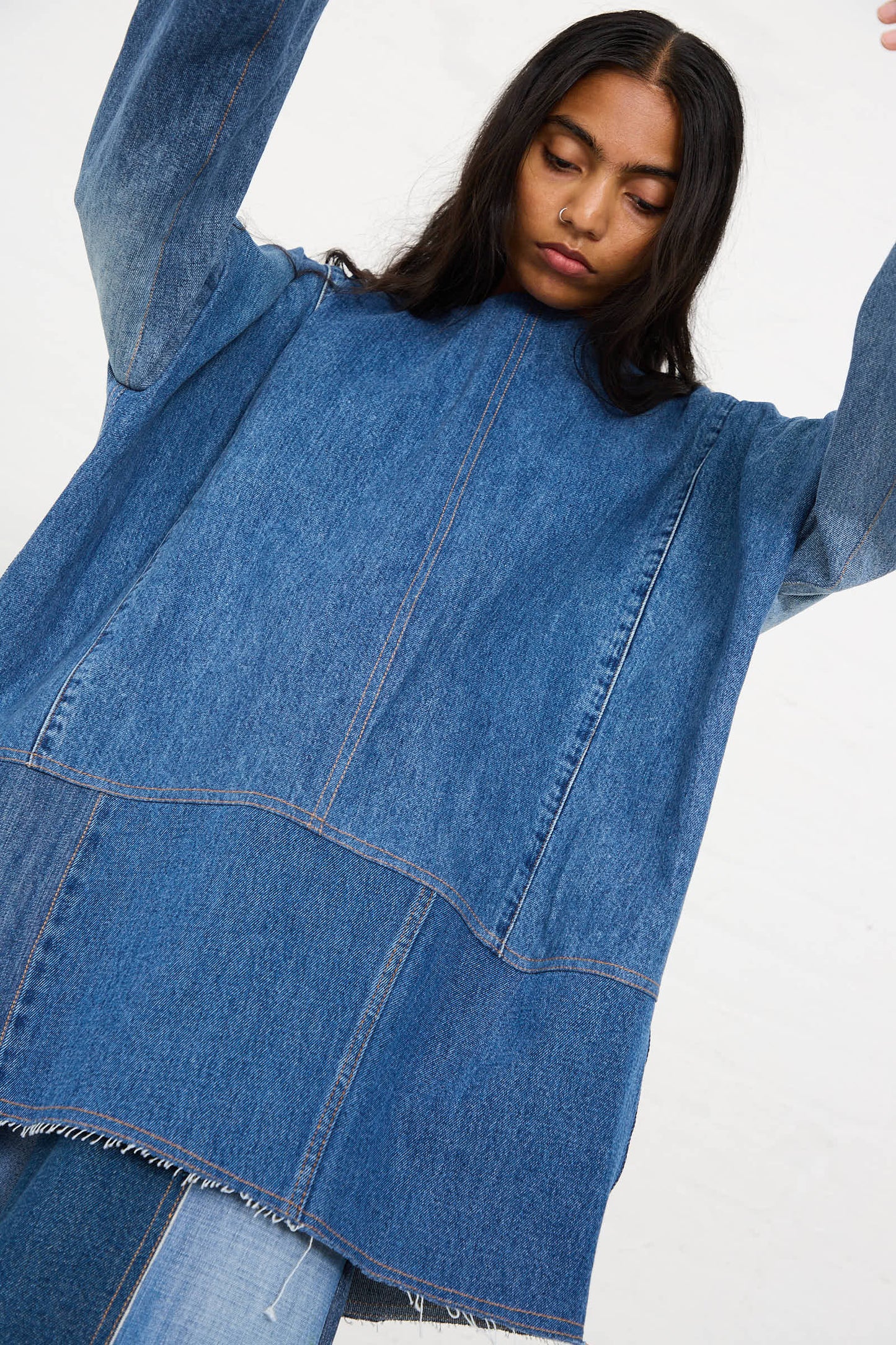 A person with long dark hair is wearing an oversized fit Salvaged Jean Patchwork Raglan Shirt in Blue by Niccolò Pasqualetti and is raising their arms against a plain white background.