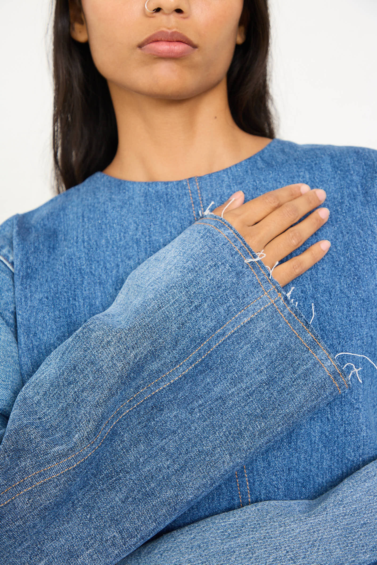 A person with long dark hair is seen from the shoulders up, wearing an oversized fit, loose-fitting Salvaged Jean Patchwork Raglan Shirt in Blue by Niccolò Pasqualetti and holding one hand over their chest.