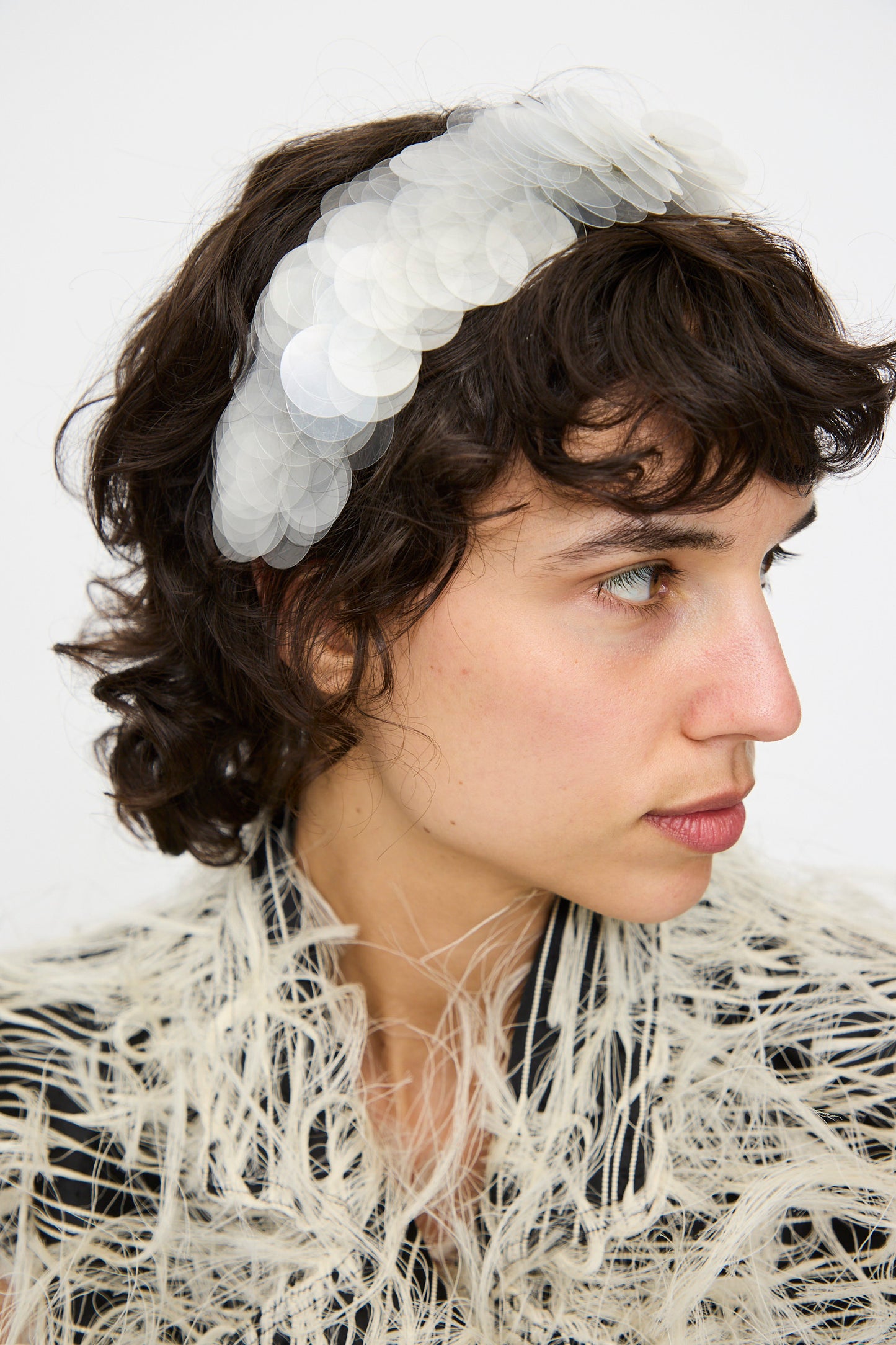 Person with short, curly brown hair wears the Sfocatura Headband in Khaki by Niccolò Pasqualetti and a textured, feathery garment, facing left against a plain background.