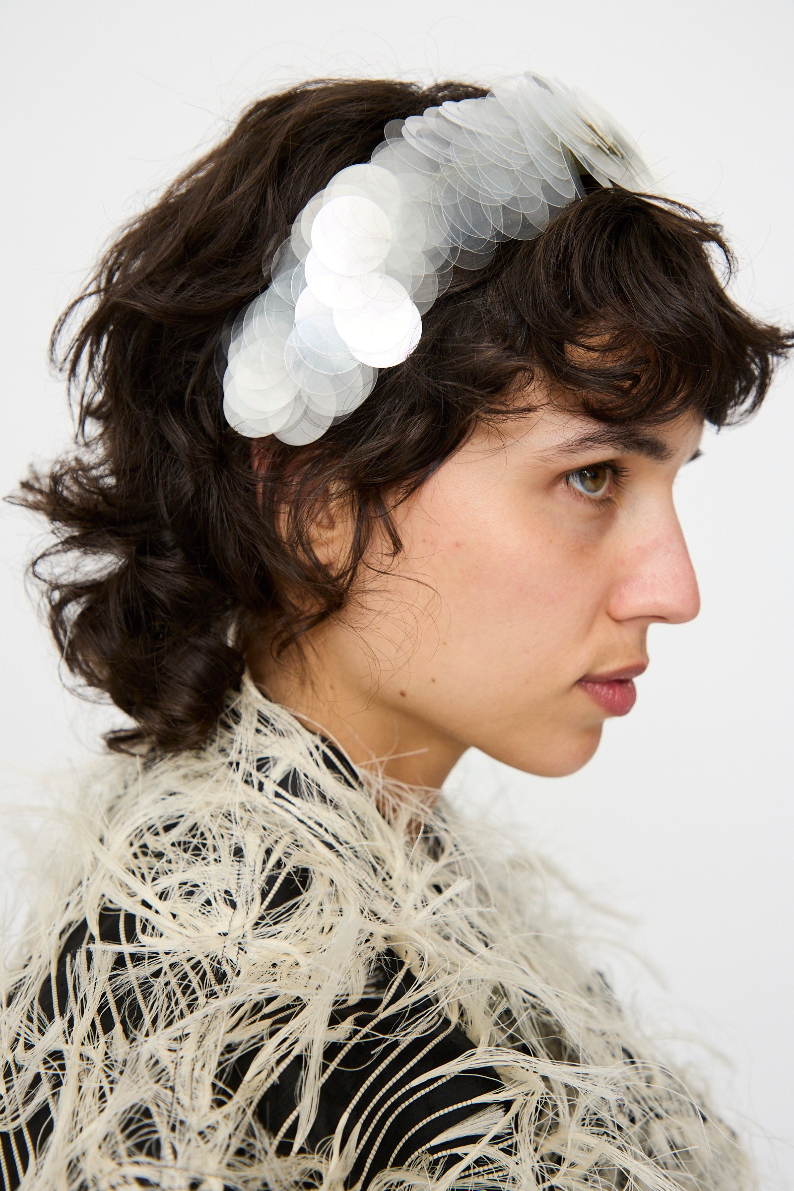 A person with short, wavy brown hair wears the Niccolò Pasqualetti Sfocatura Headband in Khaki and a feathered, textured garment.