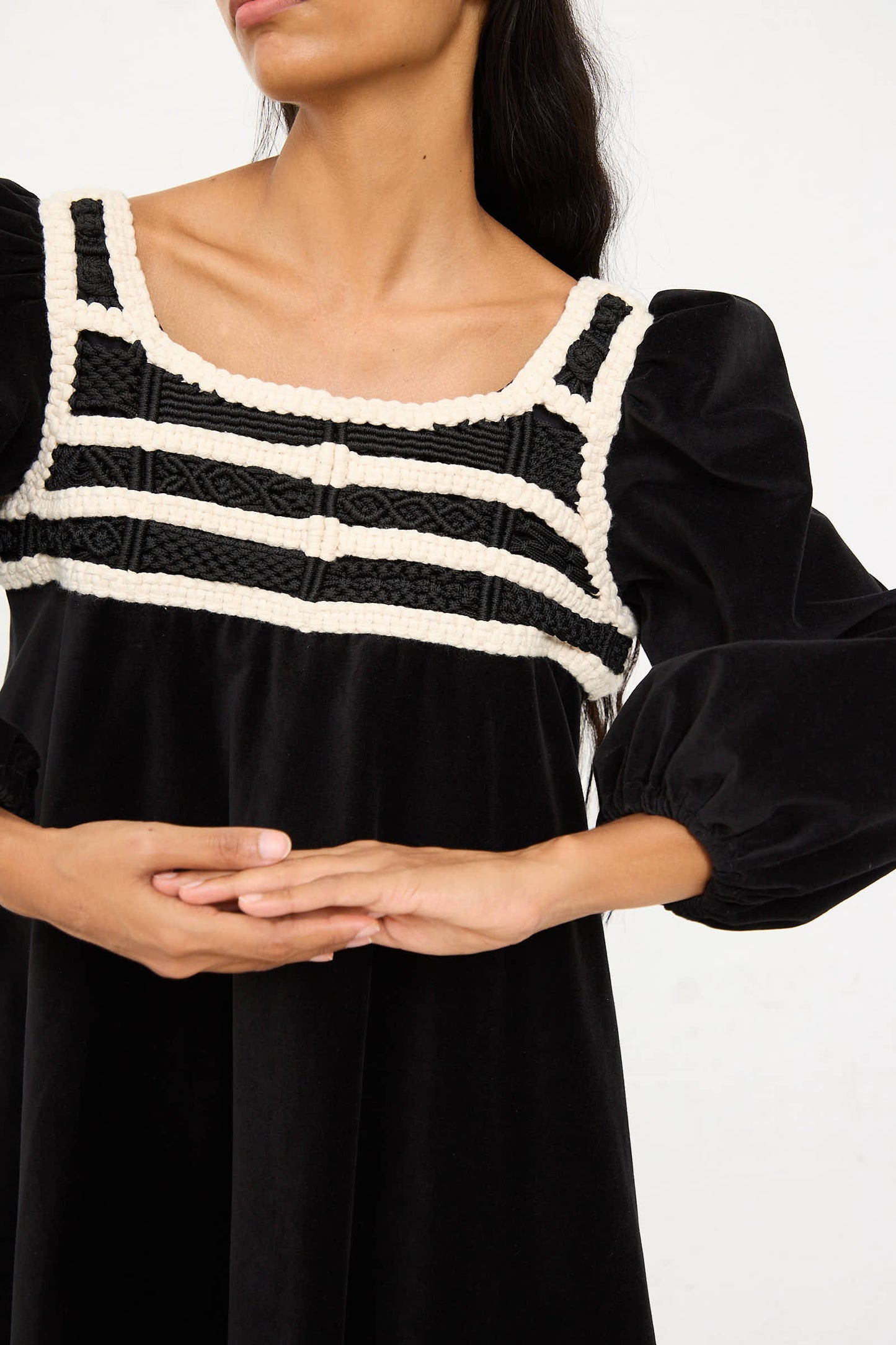 A person in the Velvet Corpetto Dress in Black by Niccolò Pasqualetti, featuring an embroidered bodice and puffed long sleeves, holds their hands together.