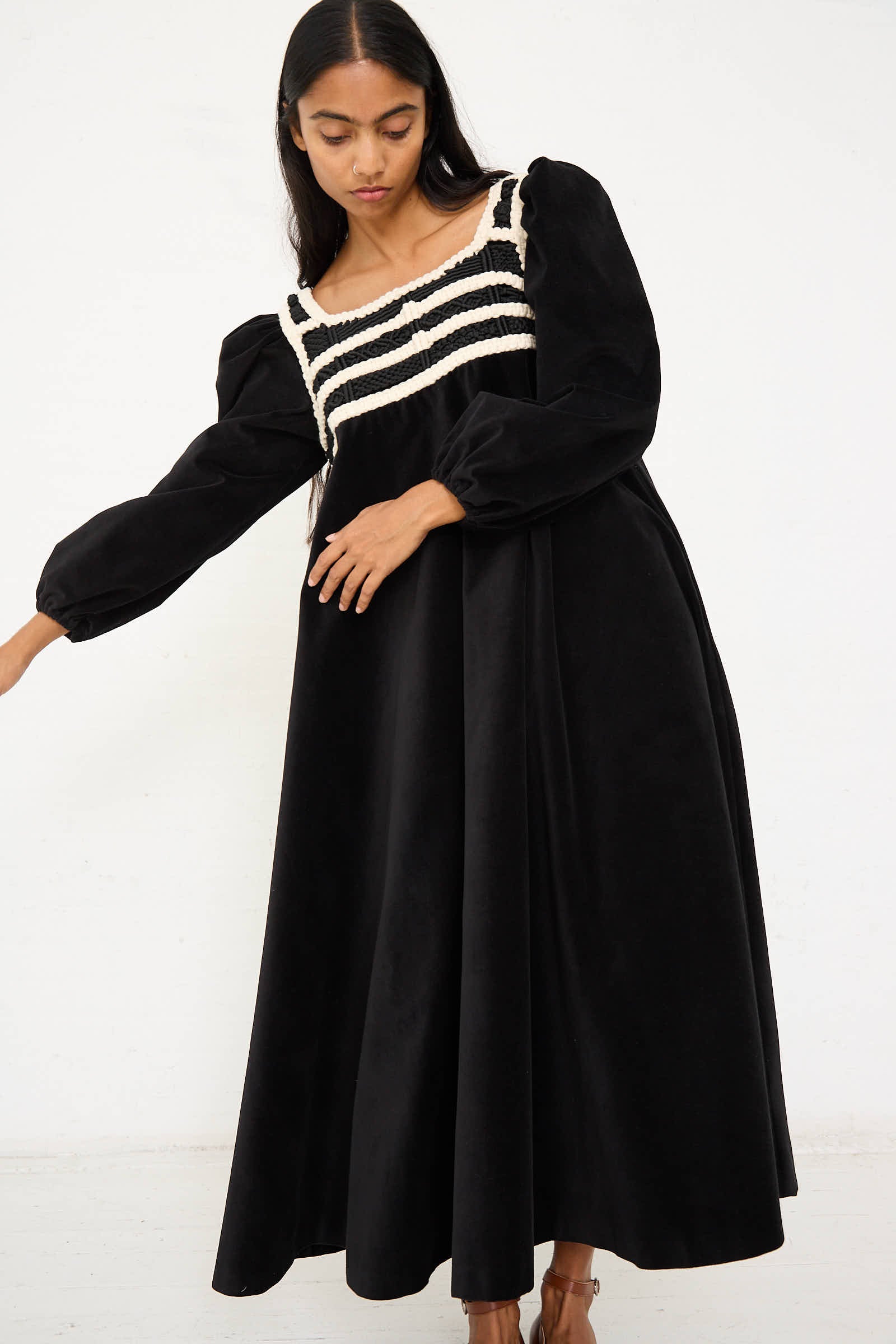 A woman with long black hair is wearing the Velvet Corpetto Dress in Black by Niccolò Pasqualetti, featuring an embroidered bodice with white and black striped detailing. She appears to be posing against a plain white background.