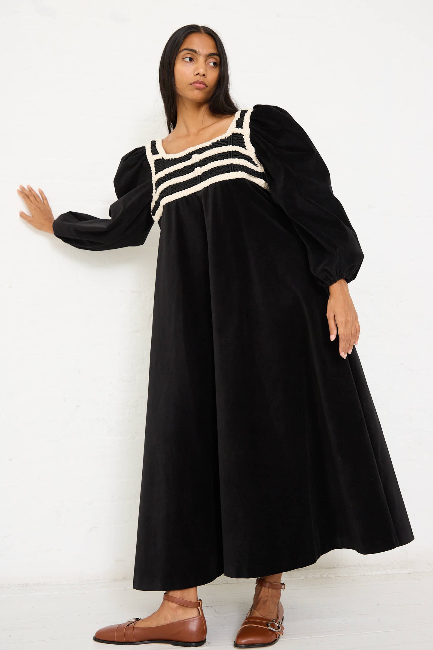 A woman stands against a white wall, wearing the Velvet Corpetto Dress in Black by Niccolò Pasqualetti, which features a striped detail across the chest, paired with brown shoes.