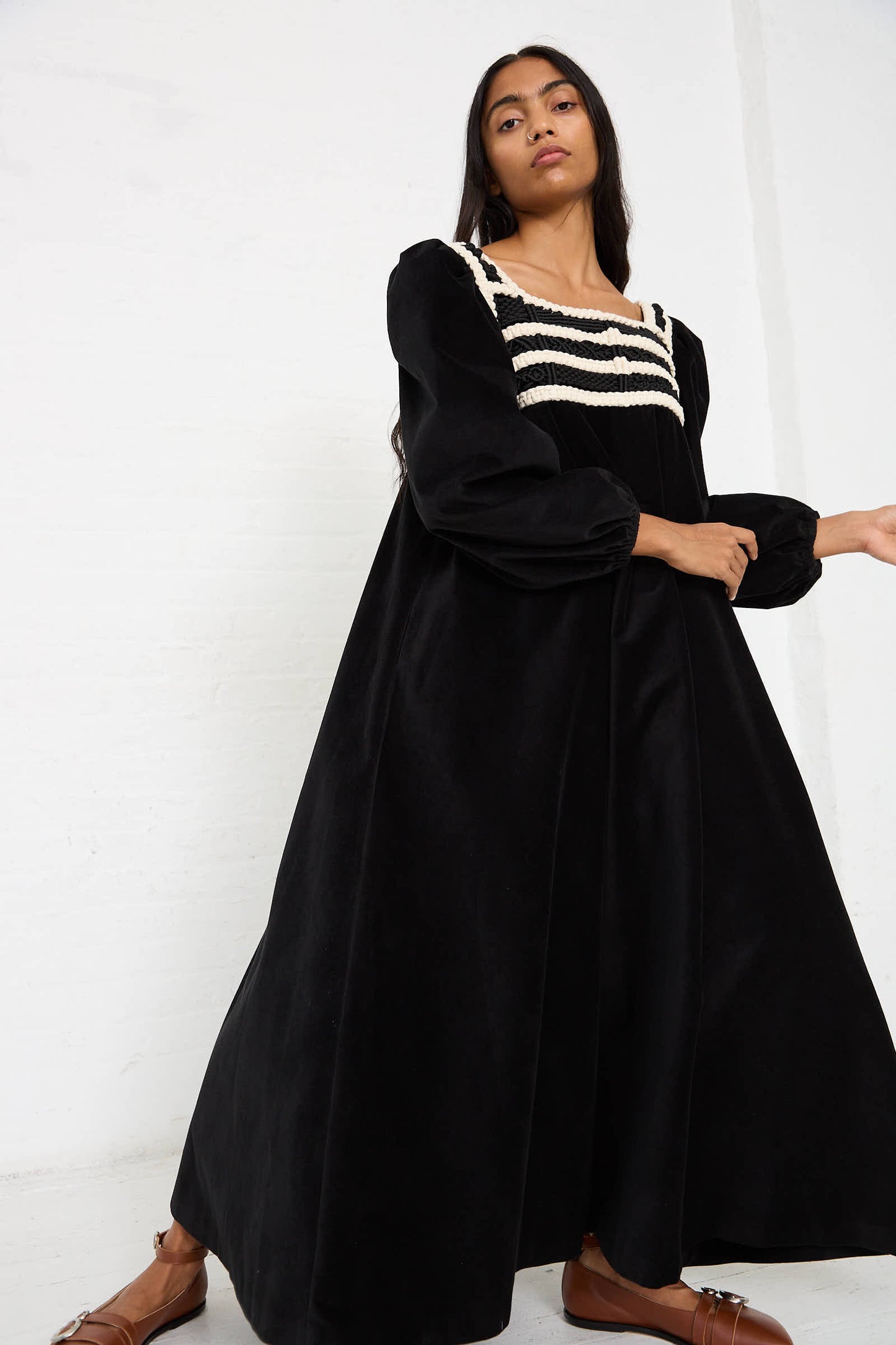 A person stands against a white background wearing the Velvet Corpetto Dress in Black by Niccolò Pasqualetti. The dress is a long, black velvet maxi with puffed sleeves and a square neckline, featuring an intricately embroidered bodice. They have long hair and are wearing brown shoes.