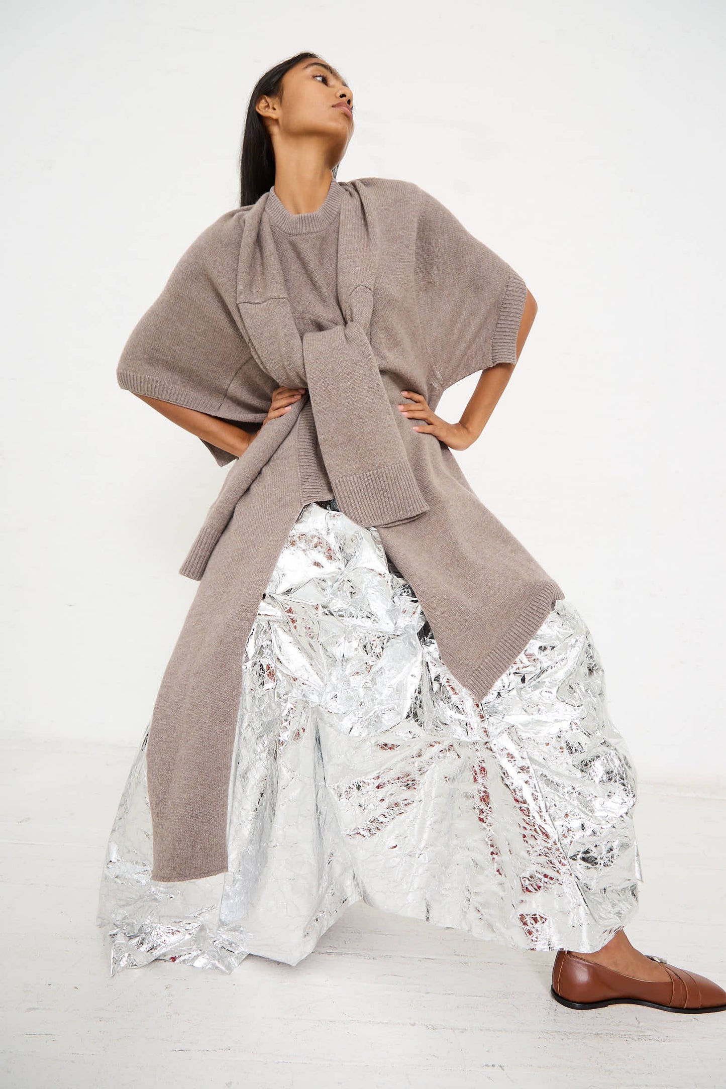 A model poses in a Niccolò Pasqualetti Wool Cashmere Blend Massa Knit Sweater in Light Brown, paired with a scarf, silver metallic oversized pants, and brown loafers against a plain white background.