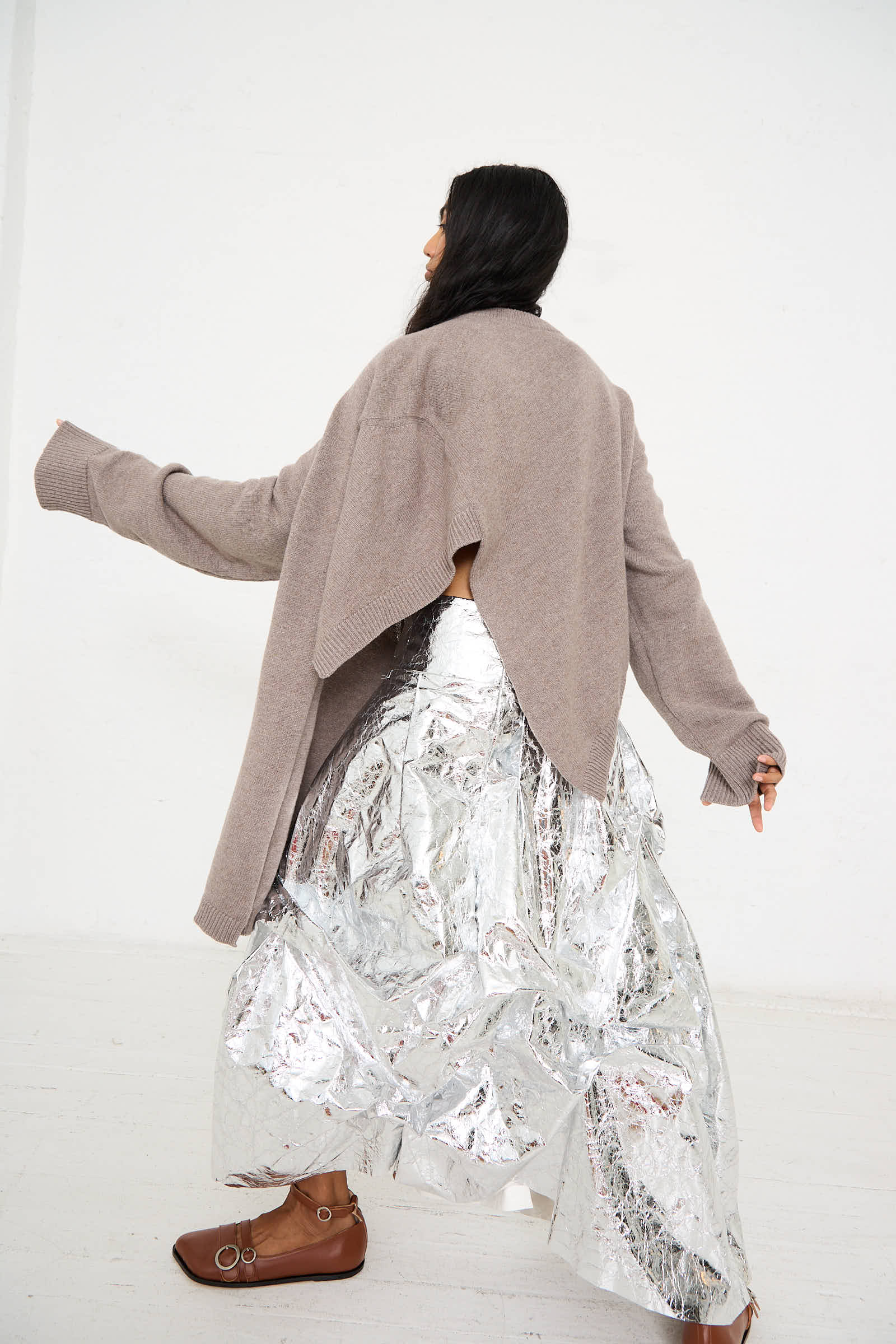 A person wearing the Niccolò Pasqualetti Wool Cashmere Blend Massa Knit Sweater in Light Brown, paired with a silver foil skirt and brown shoes, is standing against a plain white background, captured from a side-back angle.