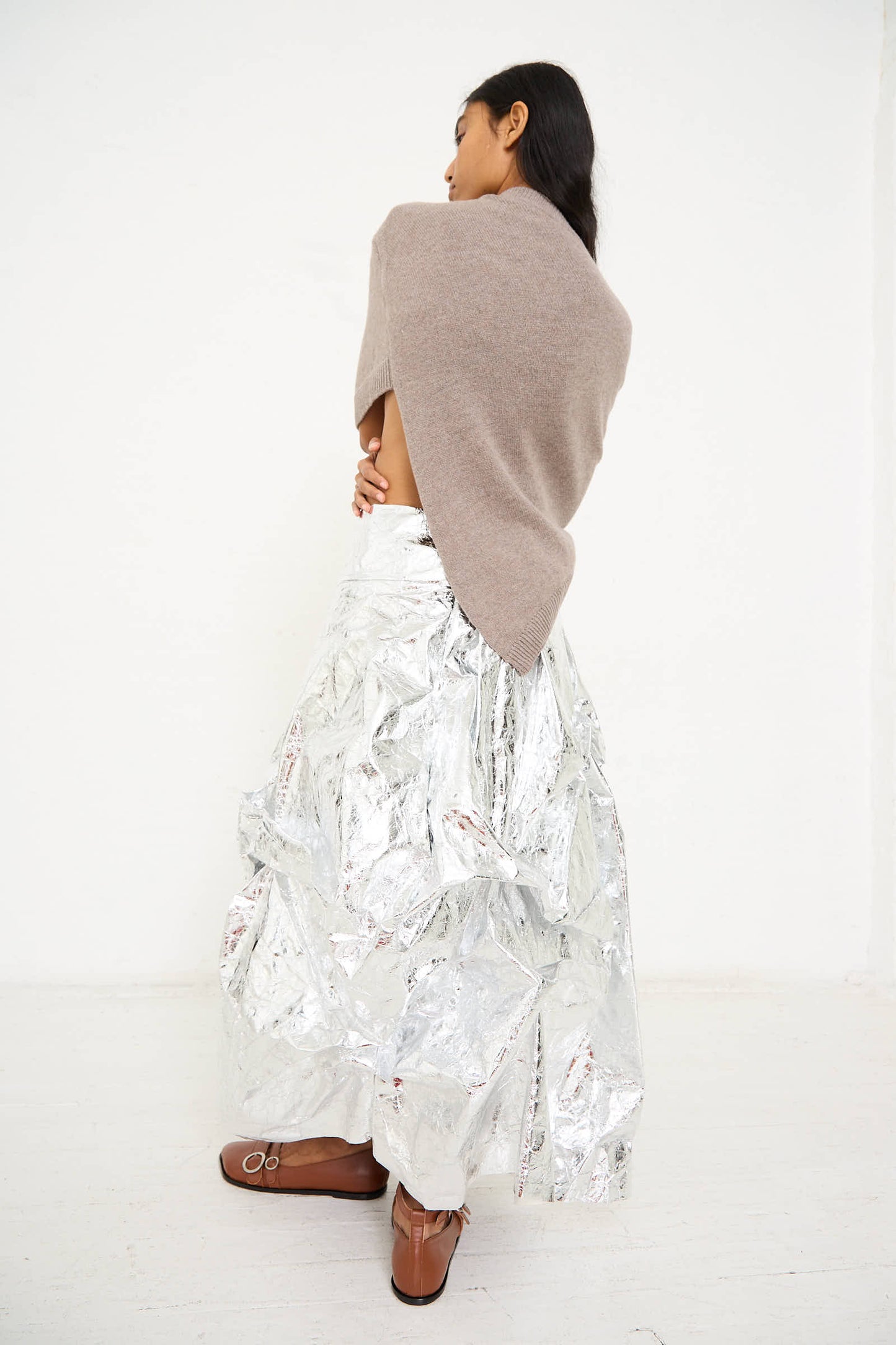 A person stands against a white background, facing away, wearing an oversized Niccolò Pasqualetti Wool Cashmere Blend Massa Knit Sweater in Light Brown draped asymmetrically over the shoulders and a shiny, silver metallic skirt paired with brown shoes.