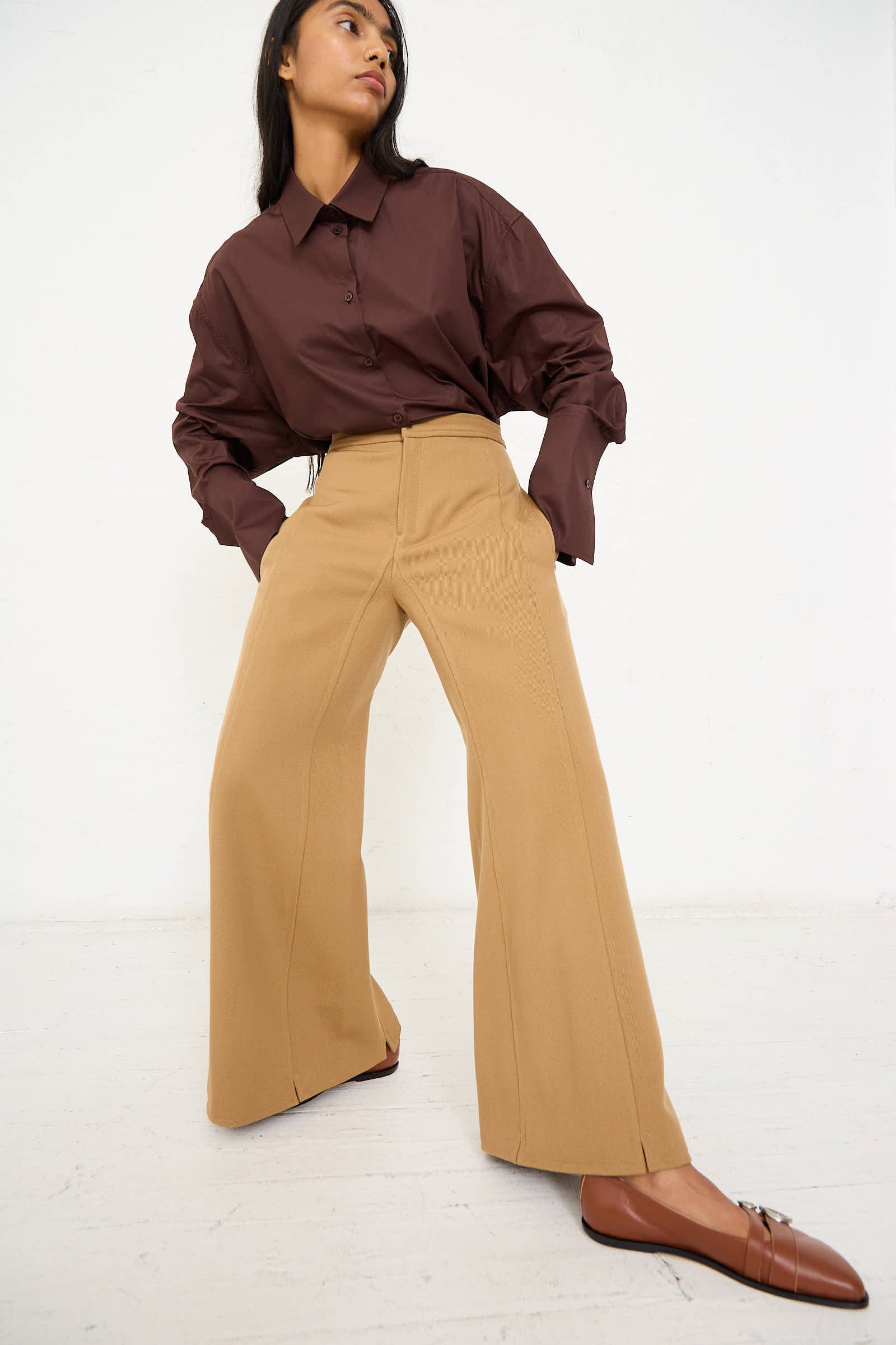 A person wearing a dark brown shirt, Niccolò Pasqualetti Wool Drill Tegola Trouser in Antique Bronze, and brown shoes stands against a white background, looking to the side.