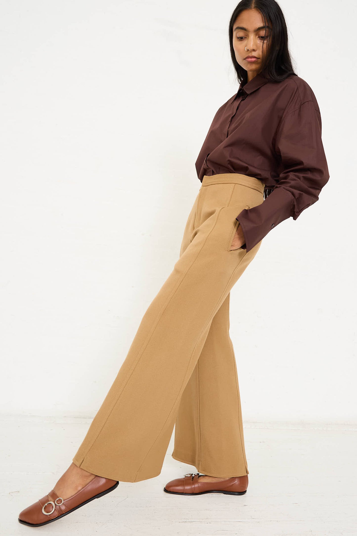 Person standing against a plain background, wearing a brown long-sleeve shirt, Niccolò Pasqualetti Wool Drill Tegola Trouser in Antique Bronze with flared legs, and brown shoes with decorative buckles.