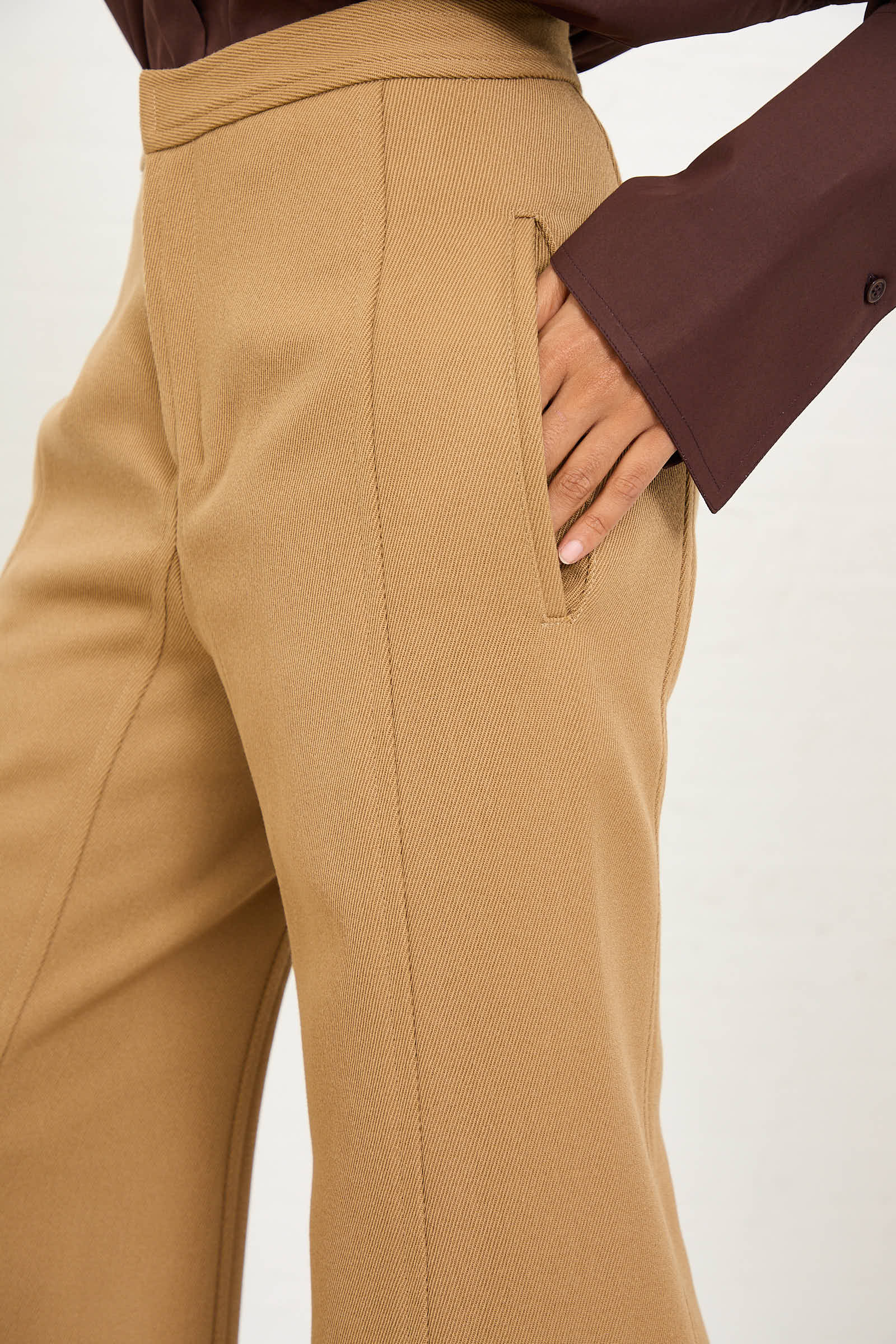 Close-up of a person wearing the Niccolò Pasqualetti Wool Drill Tegola Trouser in Antique Bronze with a hand in the right pocket. The person's shirt is long-sleeved and brown.