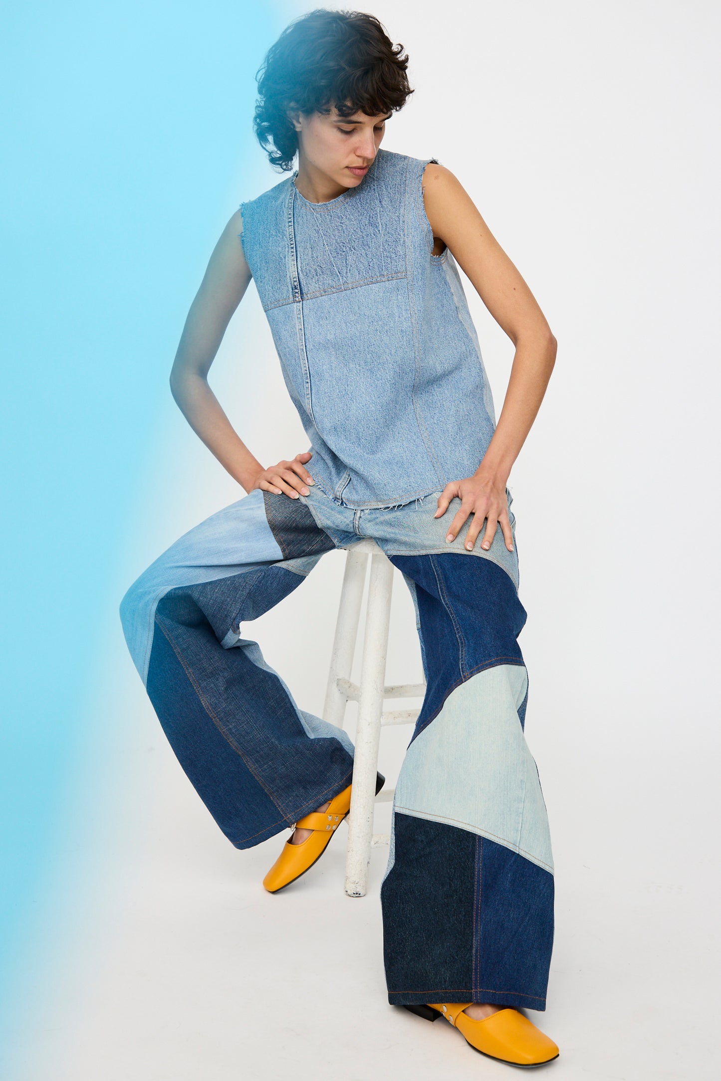 A person seated on a stool wears the Niccolò Pasqualetti Salvaged Jean Patchwork Carta Tank in Blue Denim with raw edge hems, yellow shoes, and a blue light highlighting the left side of the image.
