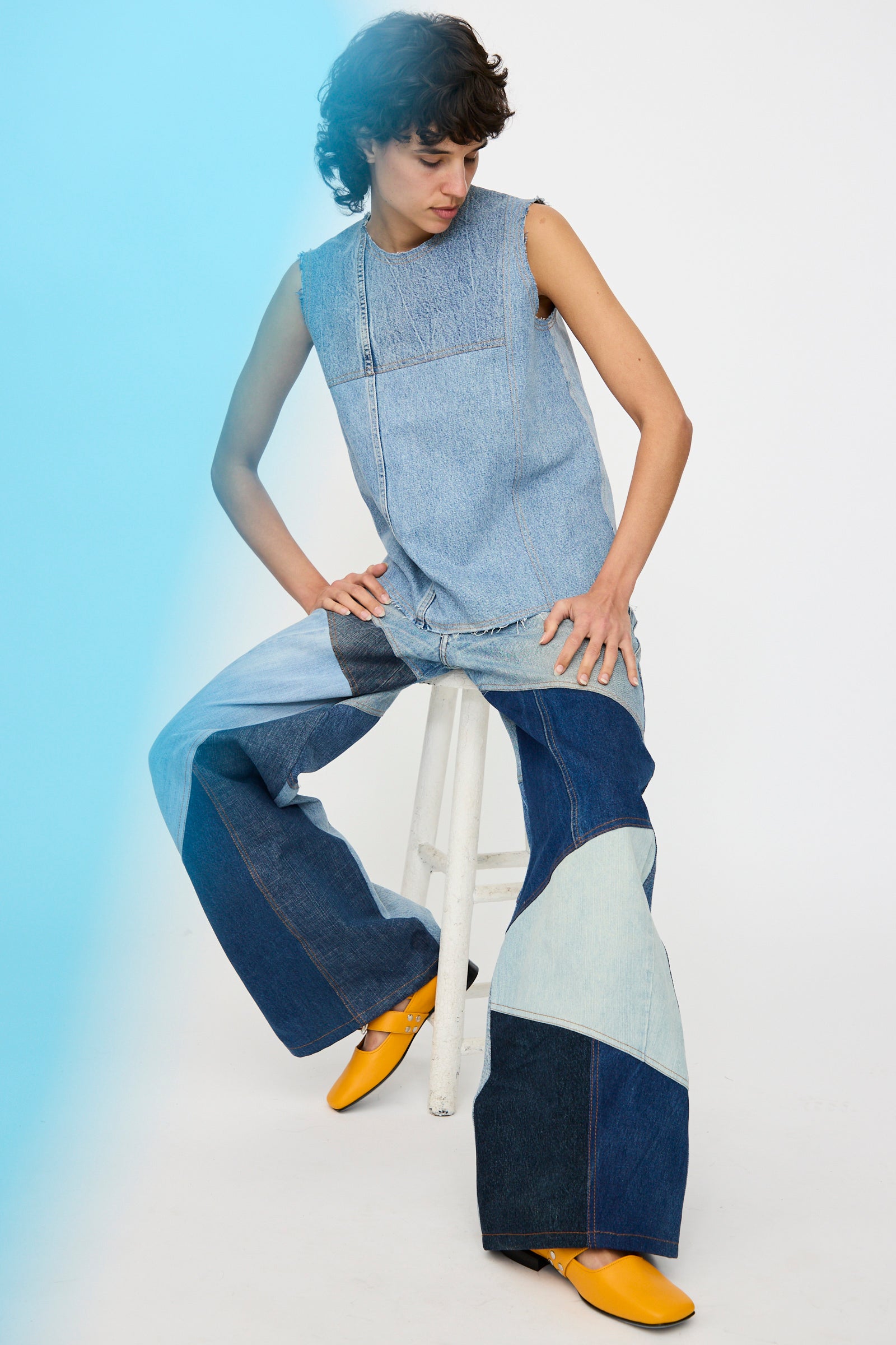 A person seated on a stool wears the Niccolò Pasqualetti Salvaged Jean Patchwork Carta Tank in Blue Denim with raw edge hems, yellow shoes, and a blue light highlighting the left side of the image.